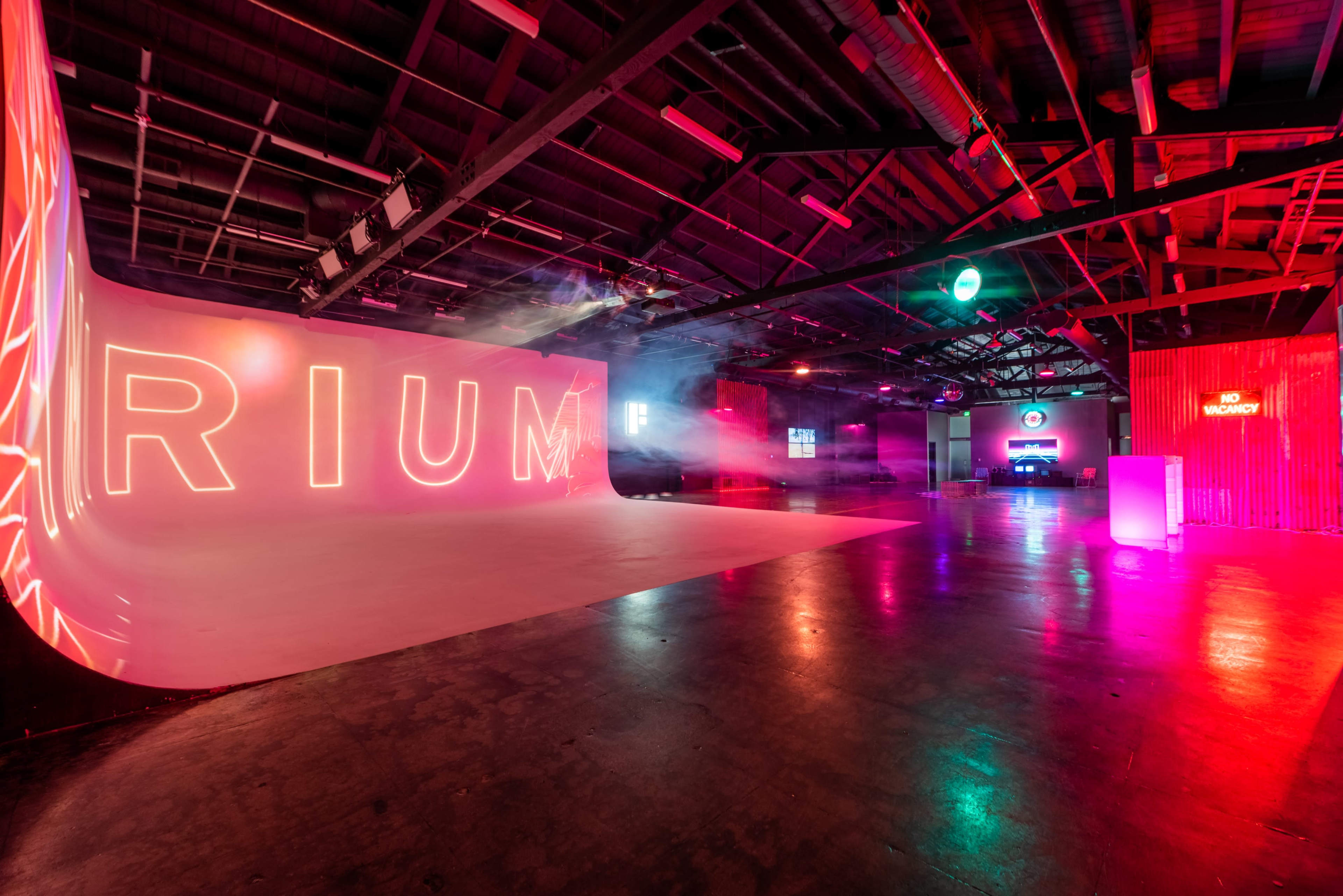 The image depicts a spacious, industrial-style venue with colorful lighting and large neon letters spelling "RIUM" on one wall.