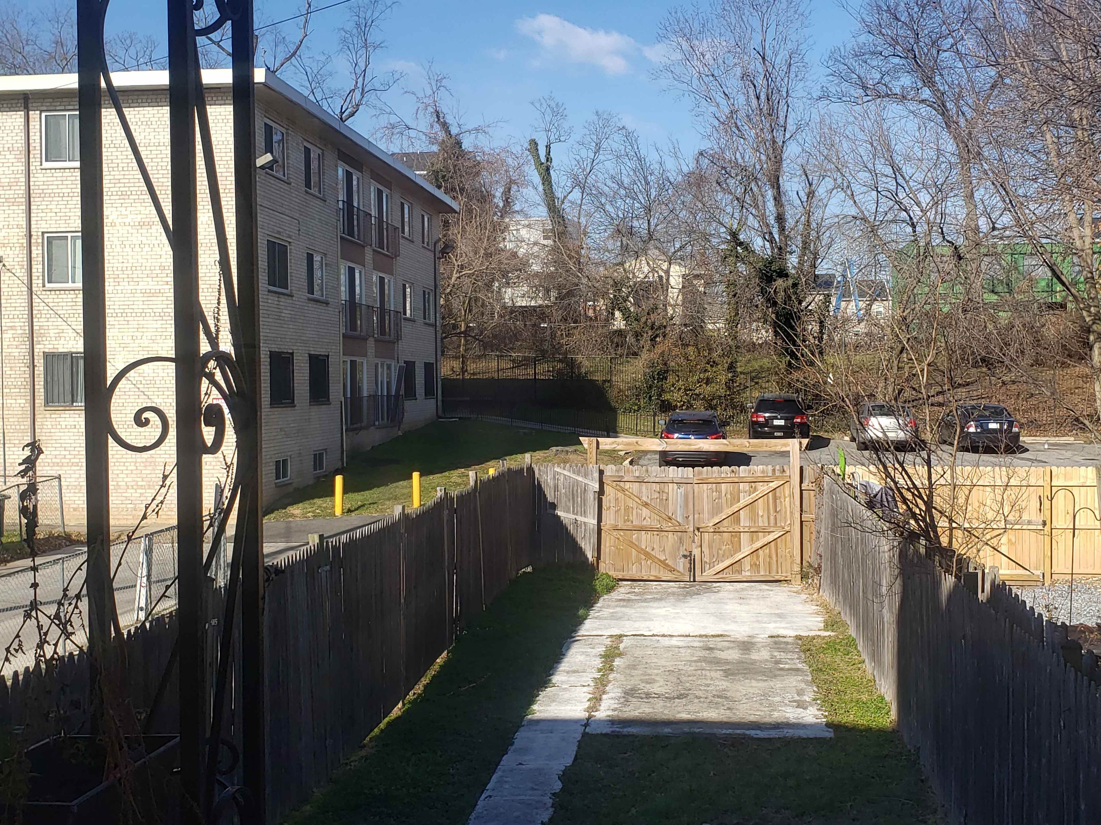 A narrow, grassy path leads to a wooden gate, bordered by a fence, with an apartment building and parking area visible in the background.