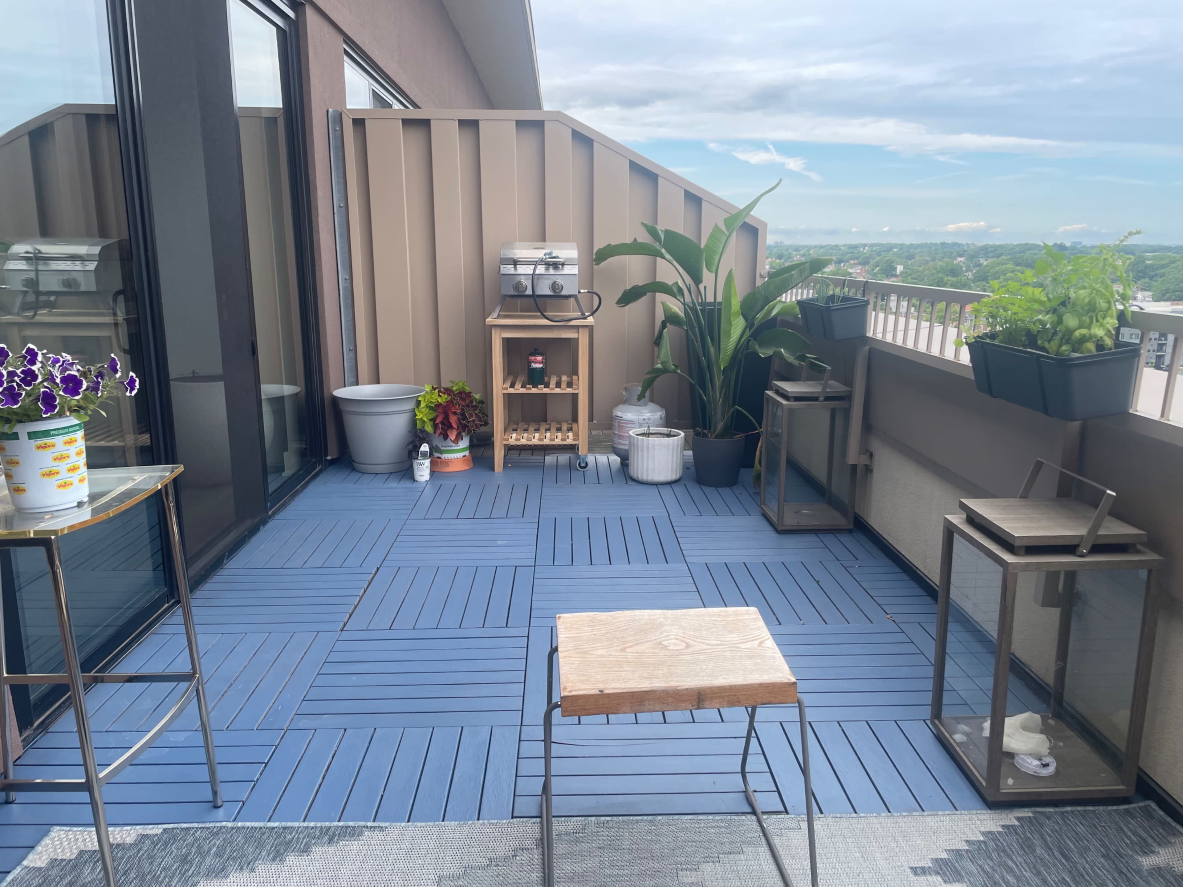 The image shows a balcony featuring outdoor furniture, potted plants, and a small grill, with a view of the sky in the background.