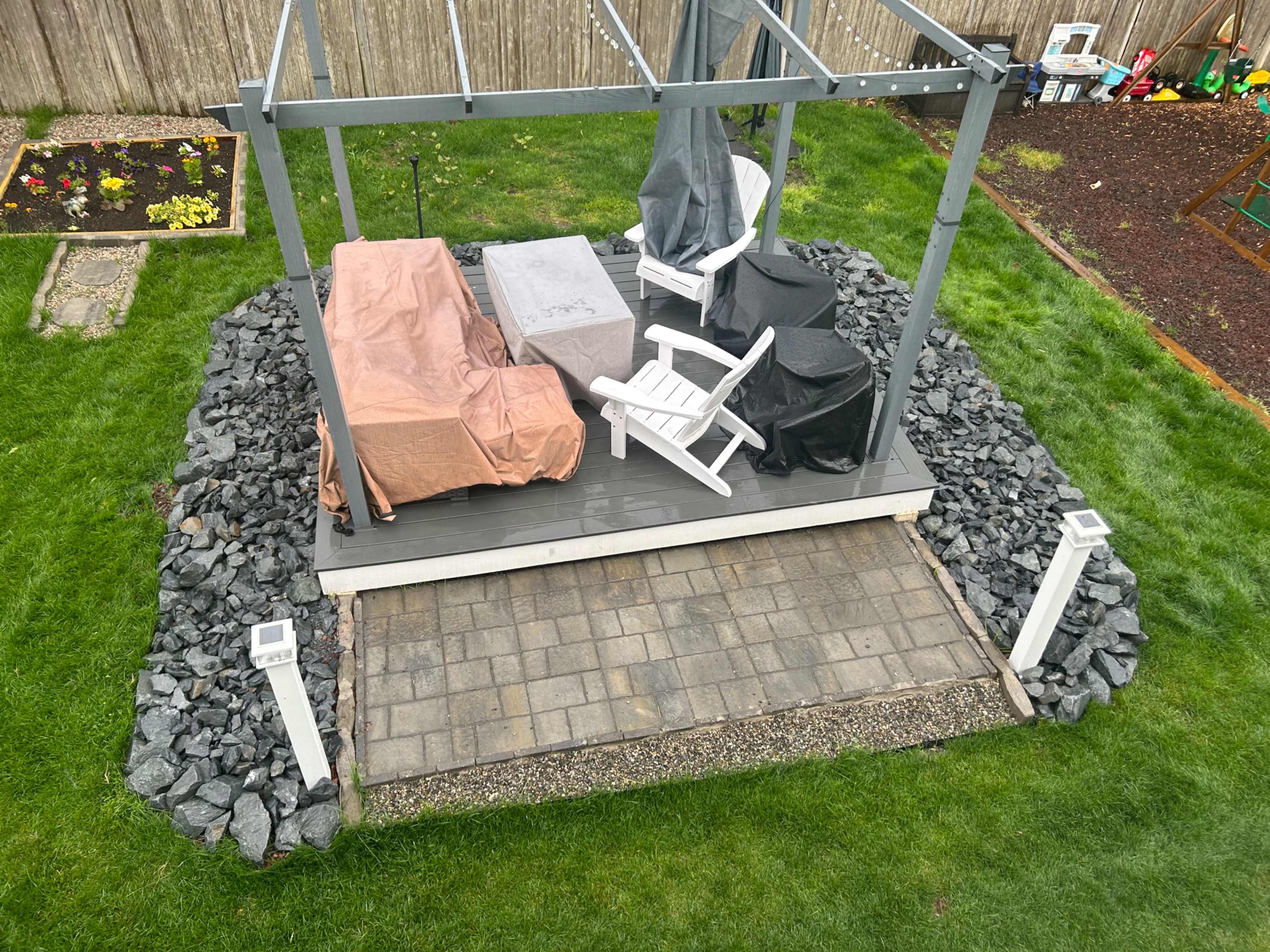 A patio with a covered seating area, featuring white chairs and a table, surrounded by gravel and grass, with a garden bed in the background.