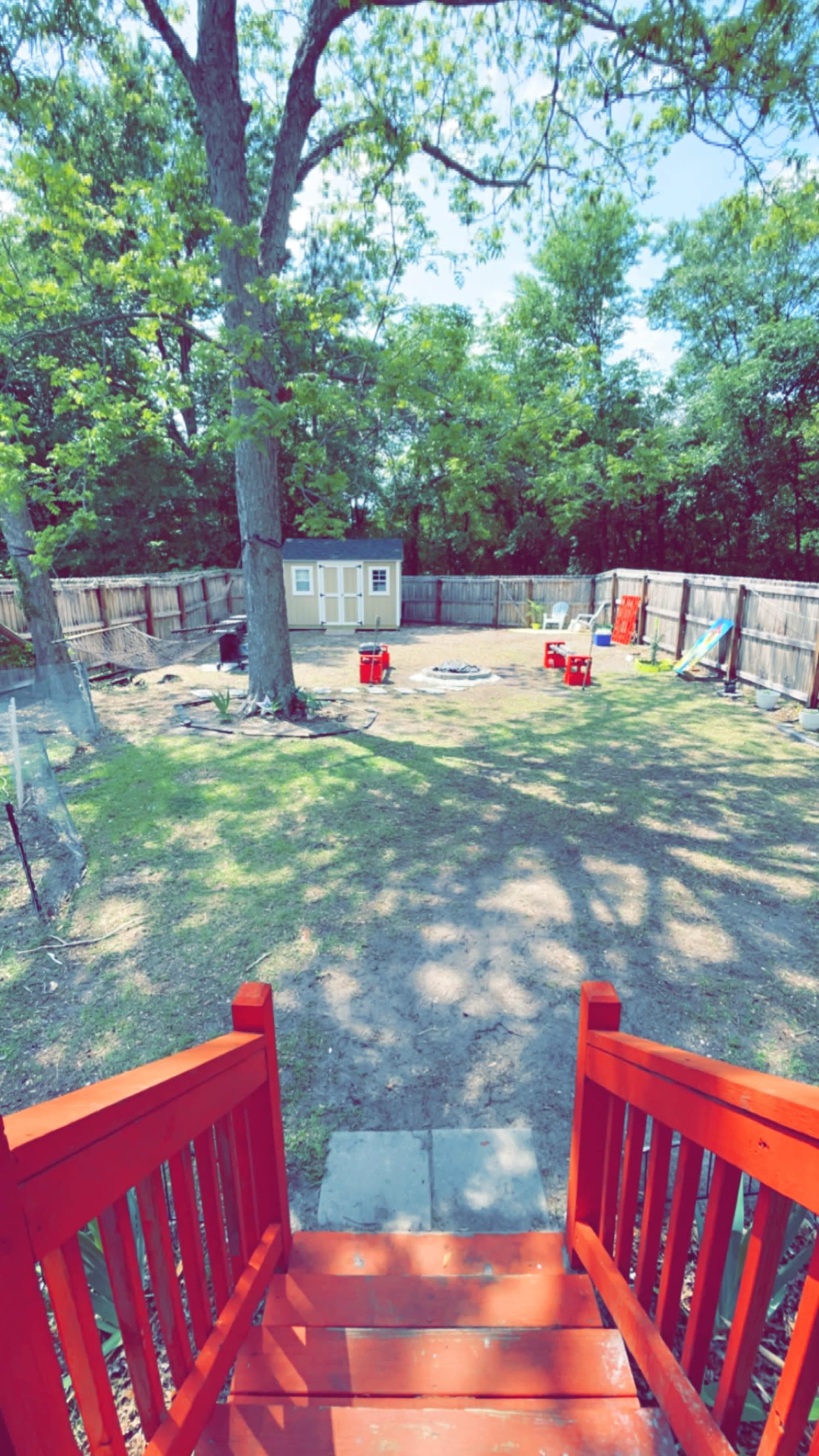 A fenced backyard features a grassy area, a shed, and red chairs arranged around a small patio.