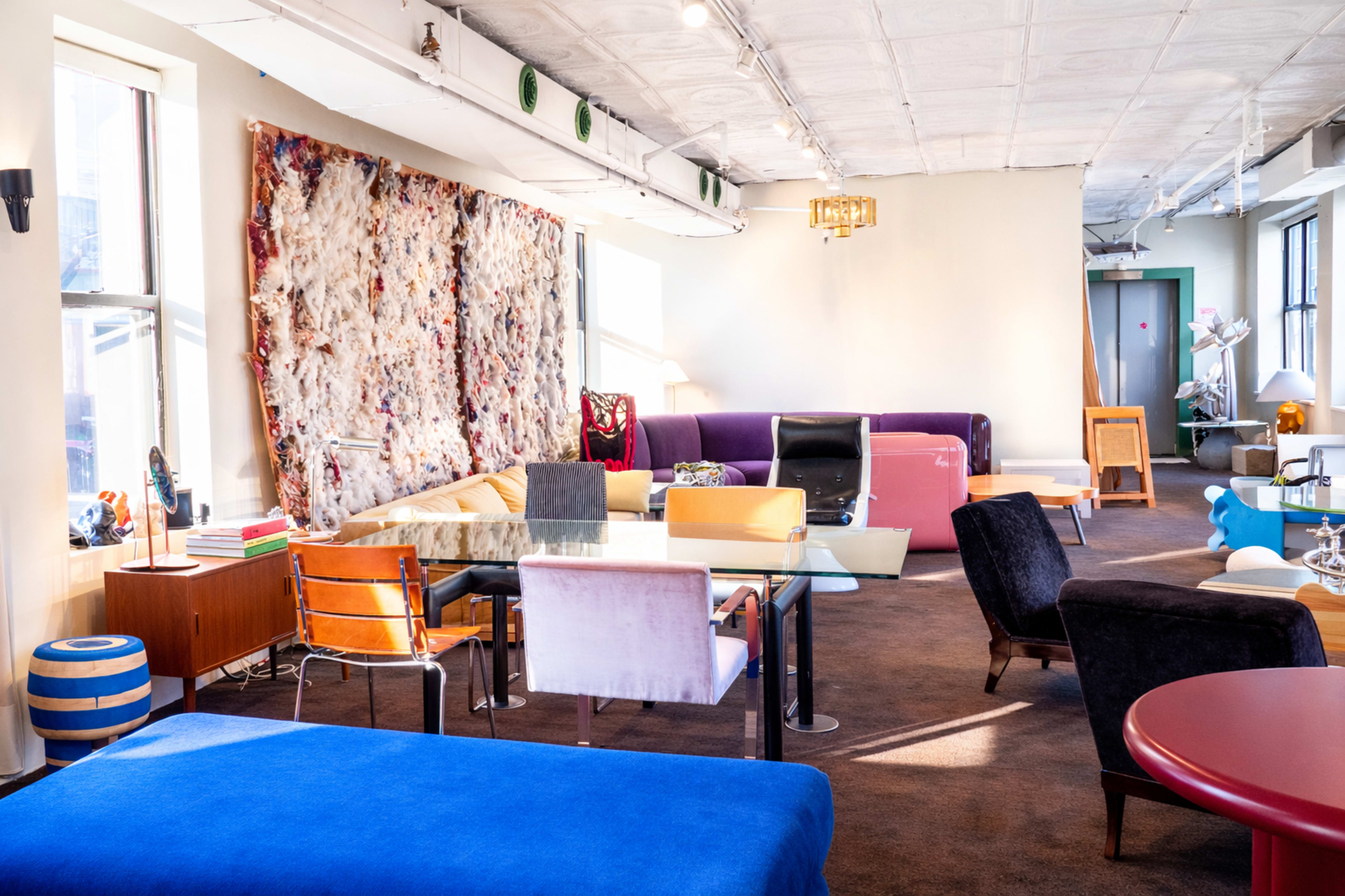 The image shows a spacious, modern lounge area furnished with various colorful chairs, a glass table, and decorative wall art.