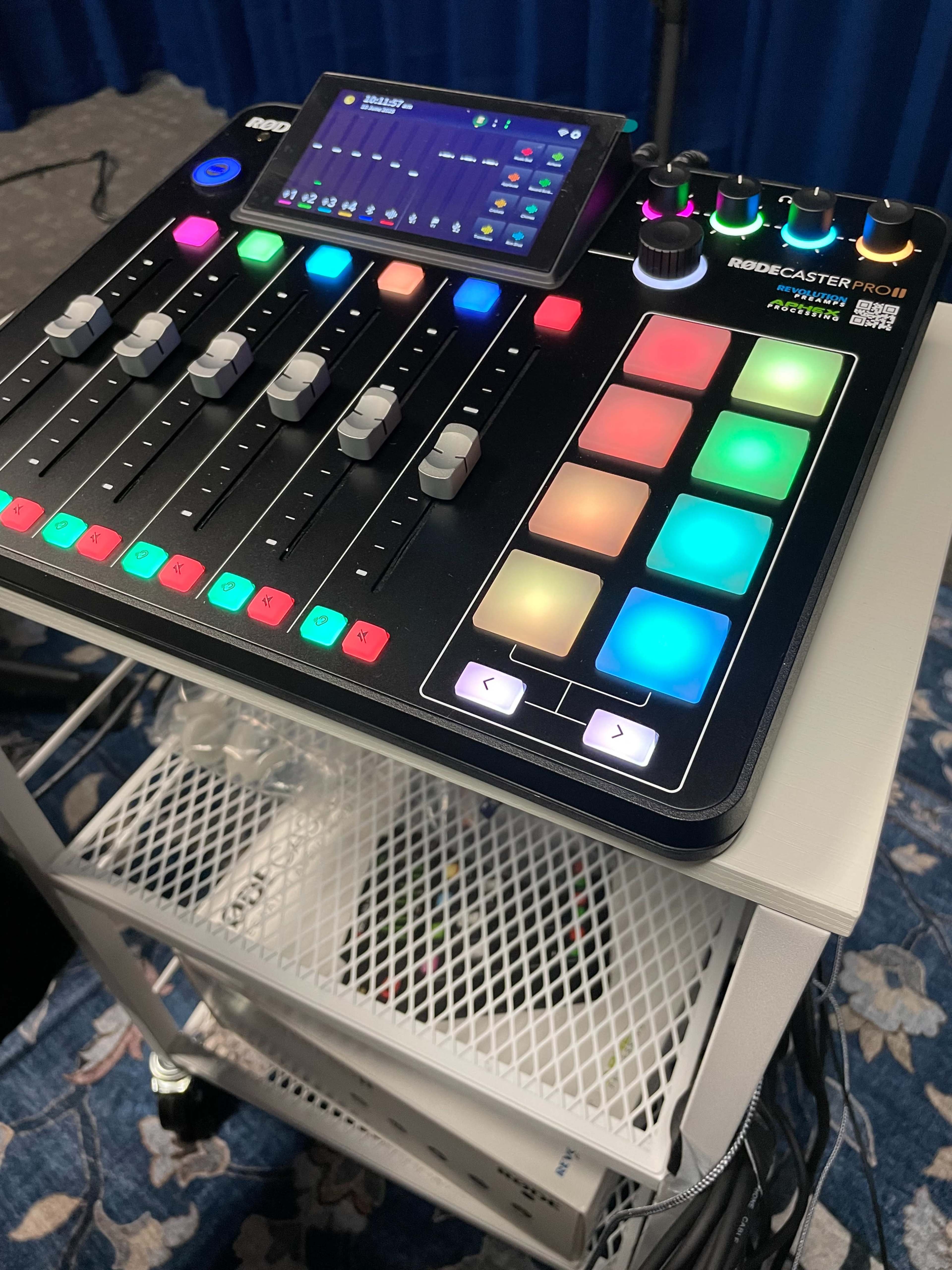 The image shows a Rodecaster Pro audio mixer with colorful buttons and faders, placed on a cart with a white mesh shelf below it.