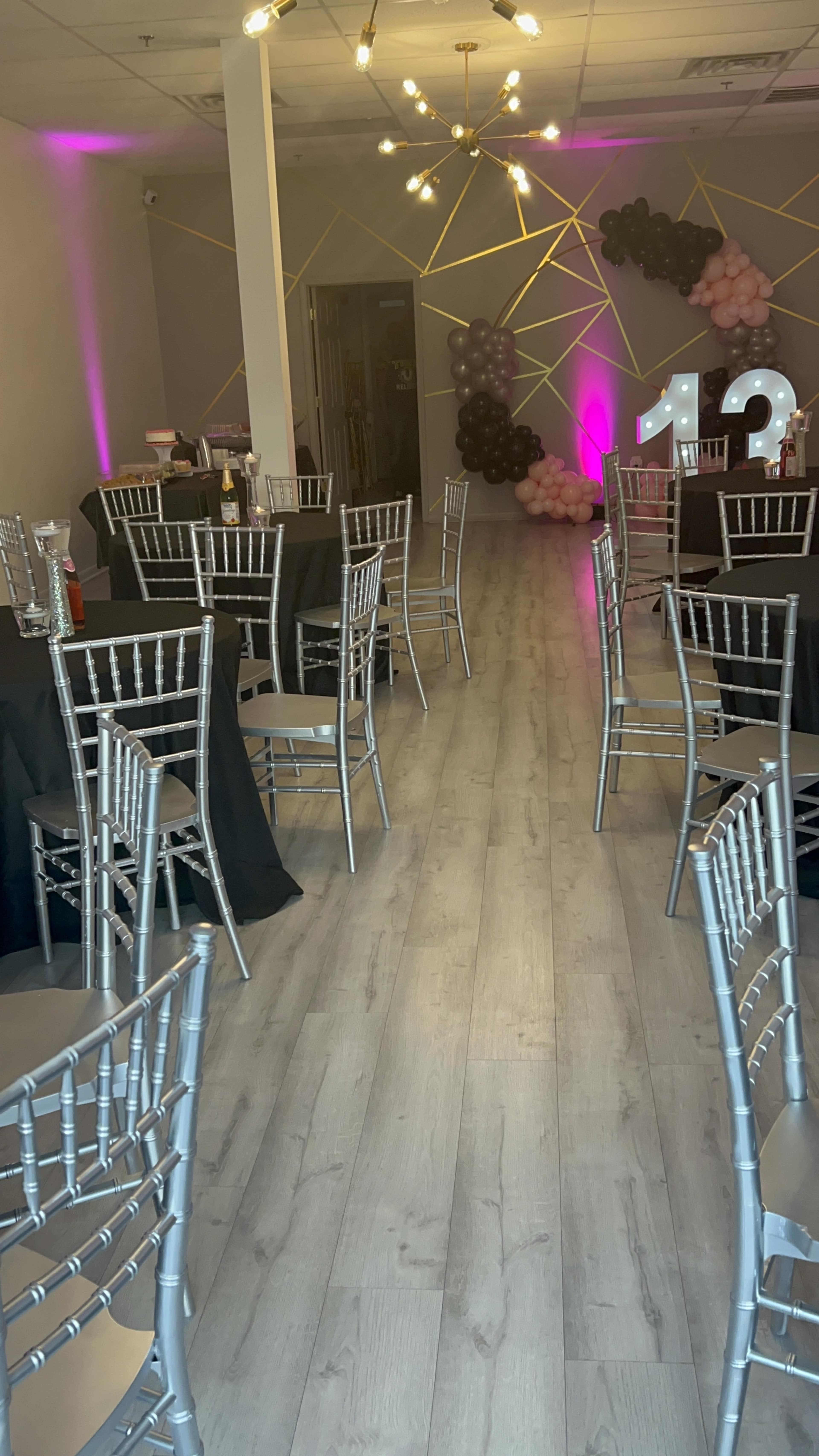 The image shows a party venue with silver chiavari chairs and black tablecloths arranged in rows, featuring a balloon display and a large illuminated "13" at the back.
