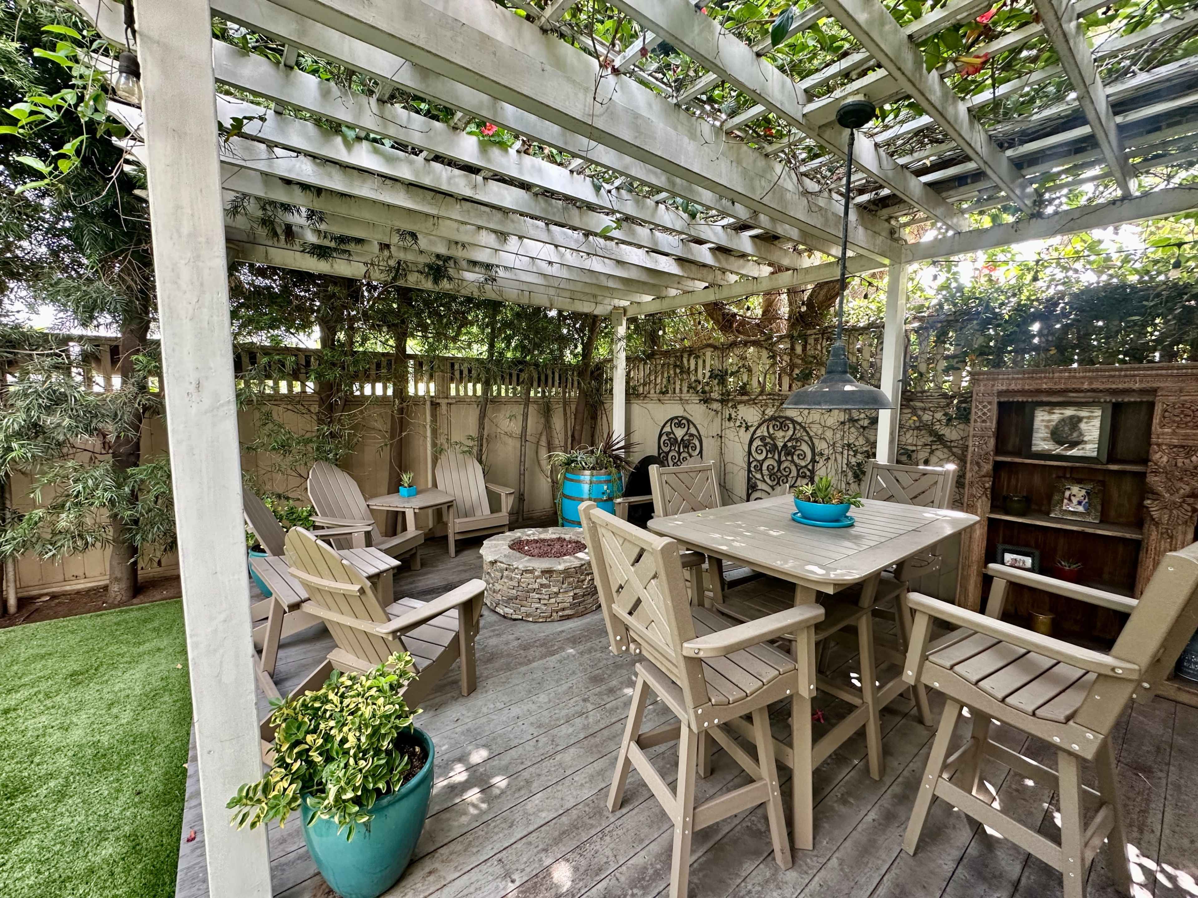 The image shows a covered outdoor seating area with a wooden table, chairs, and a stone fire pit surrounded by greenery.
