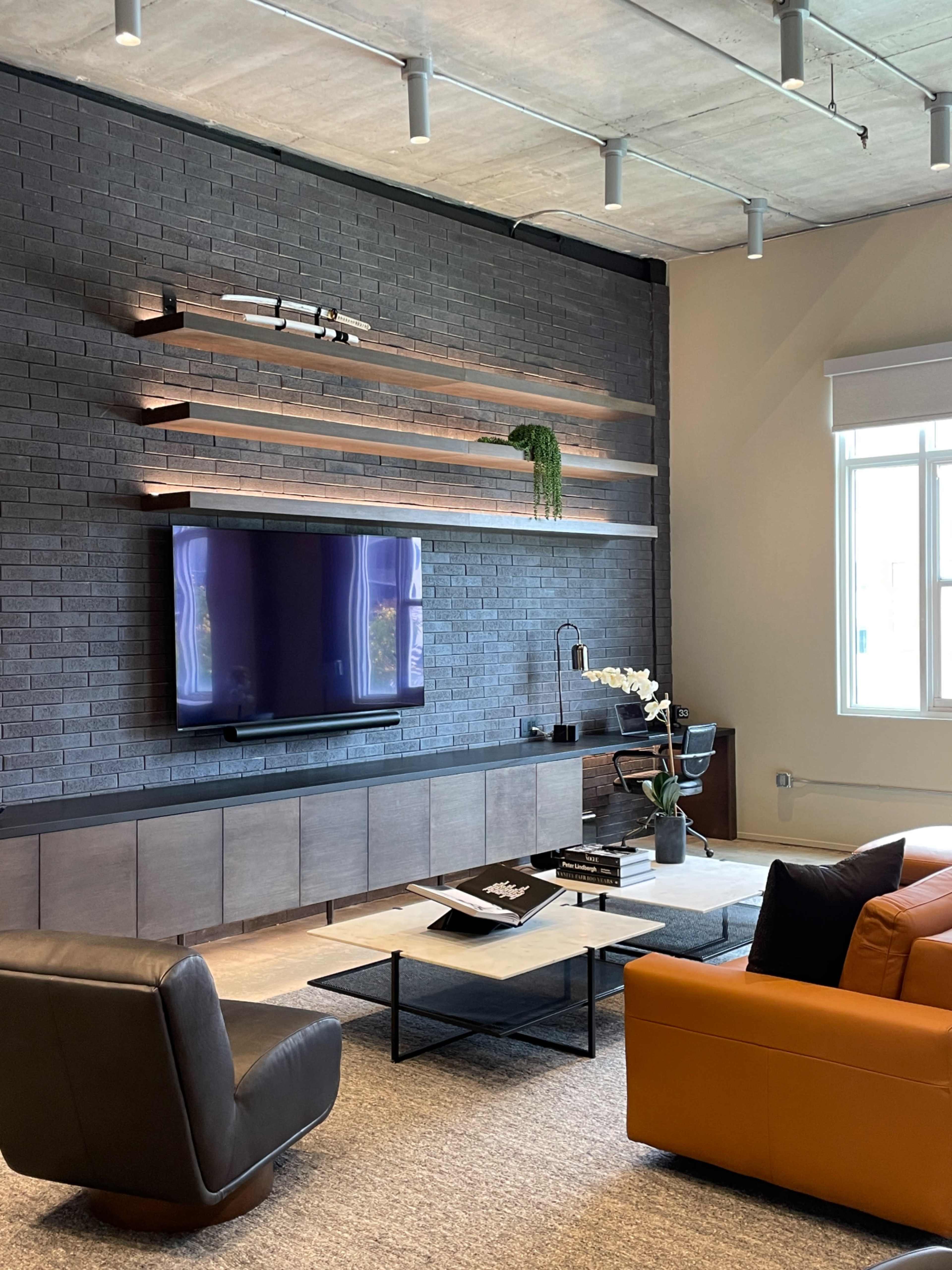The living room features a mounted television on a black brick wall with wooden shelves, an orange sofa, and a sliding glass door that allows natural light.