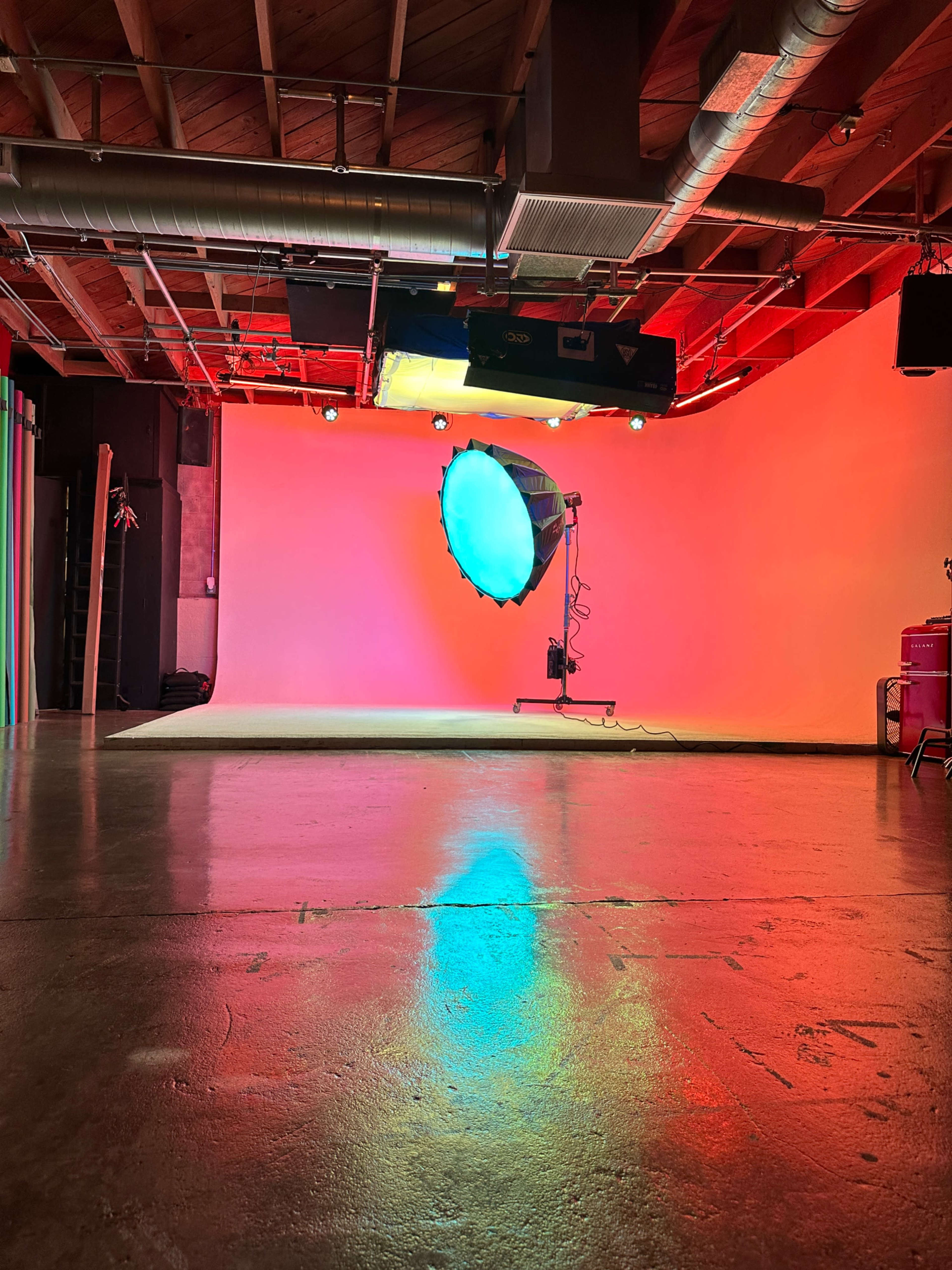 The image shows a photography studio with a brightly lit backdrop and a large spotlight casting colorful reflections on the polished floor.