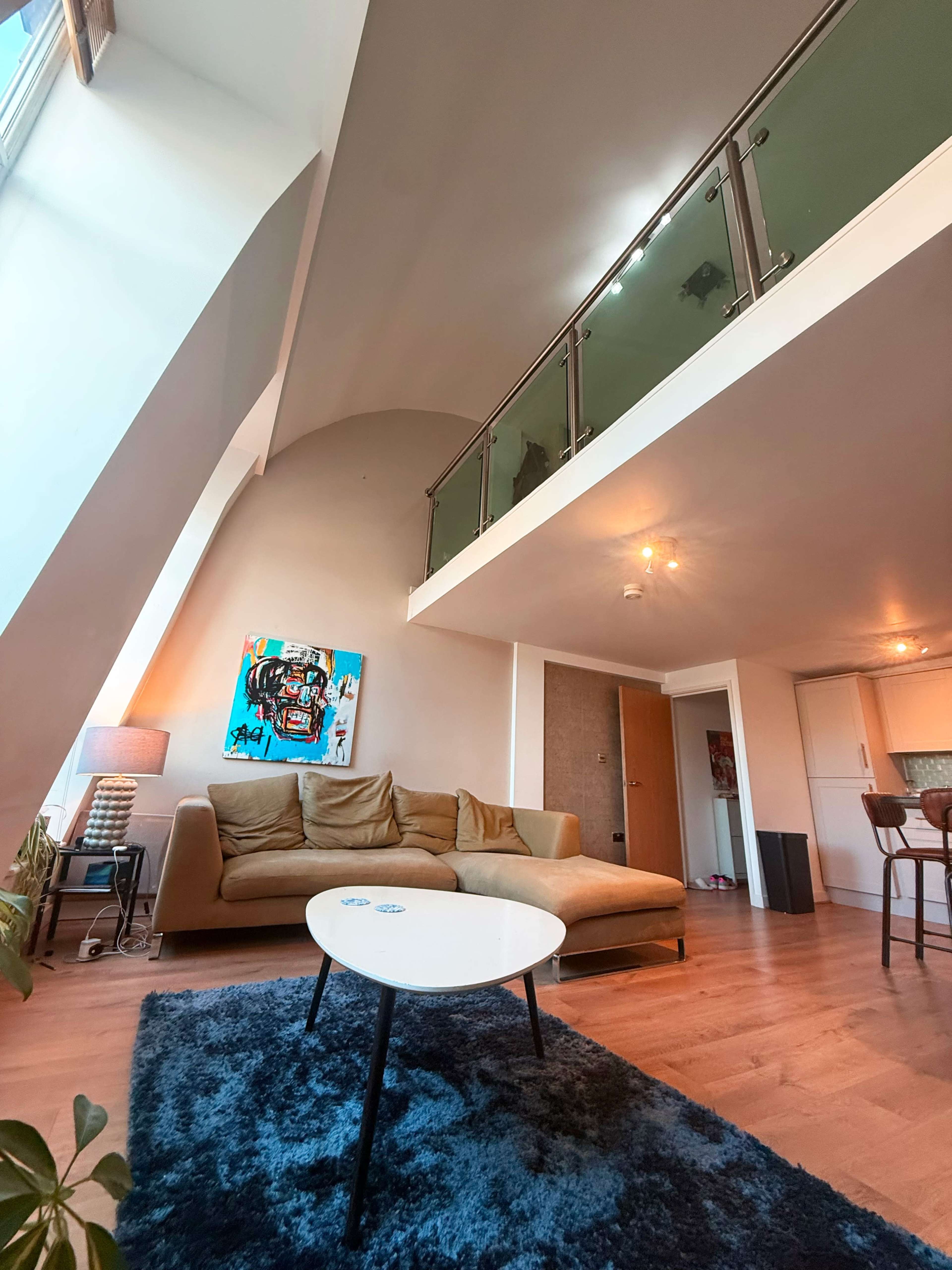 The image shows a modern living room with a brown sofa, a white coffee table, and a large artwork on the wall, overlooking a second-level balcony.