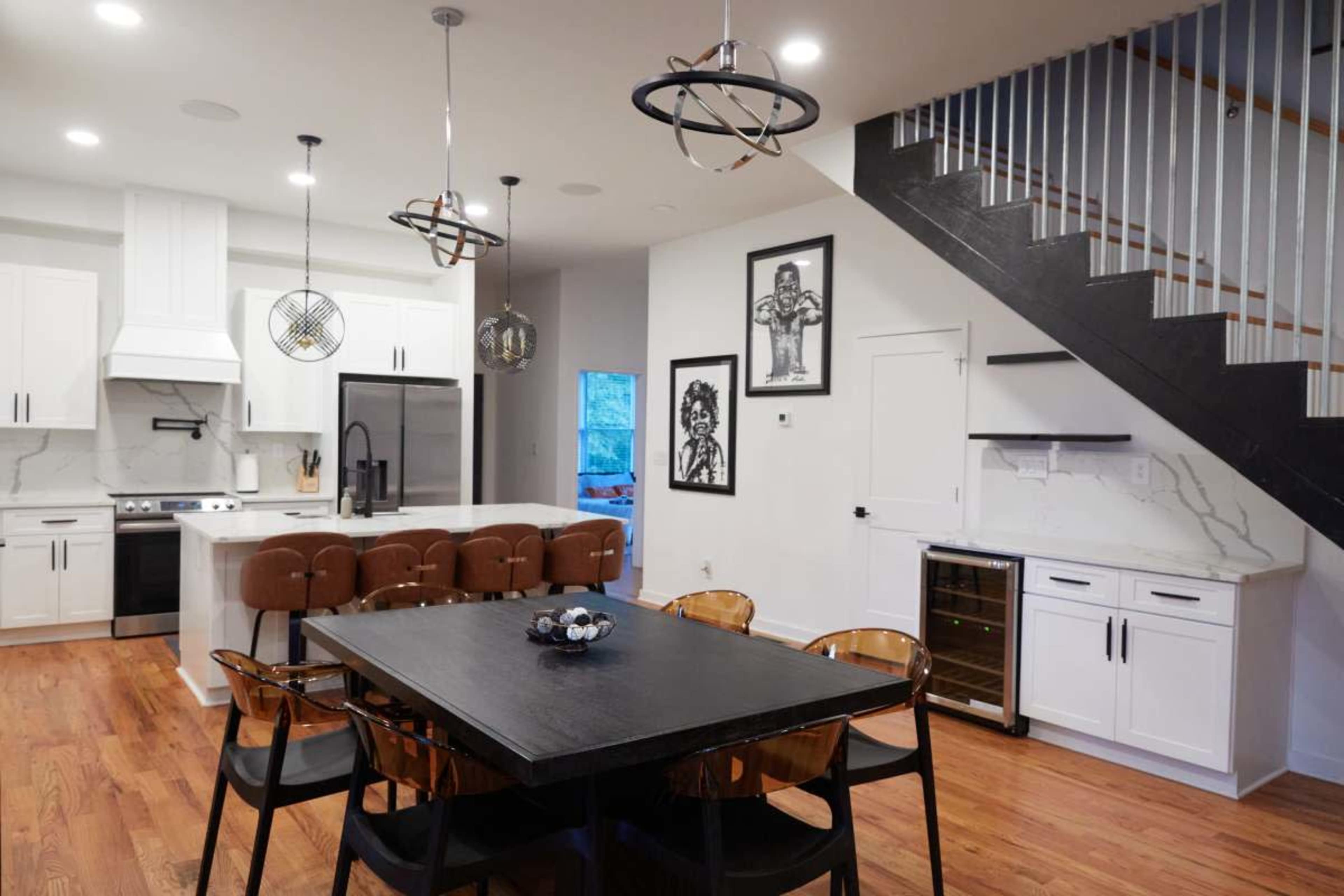 The image shows a modern kitchen and dining area with a black table, brown chairs, white cabinets, and a staircase in the background.