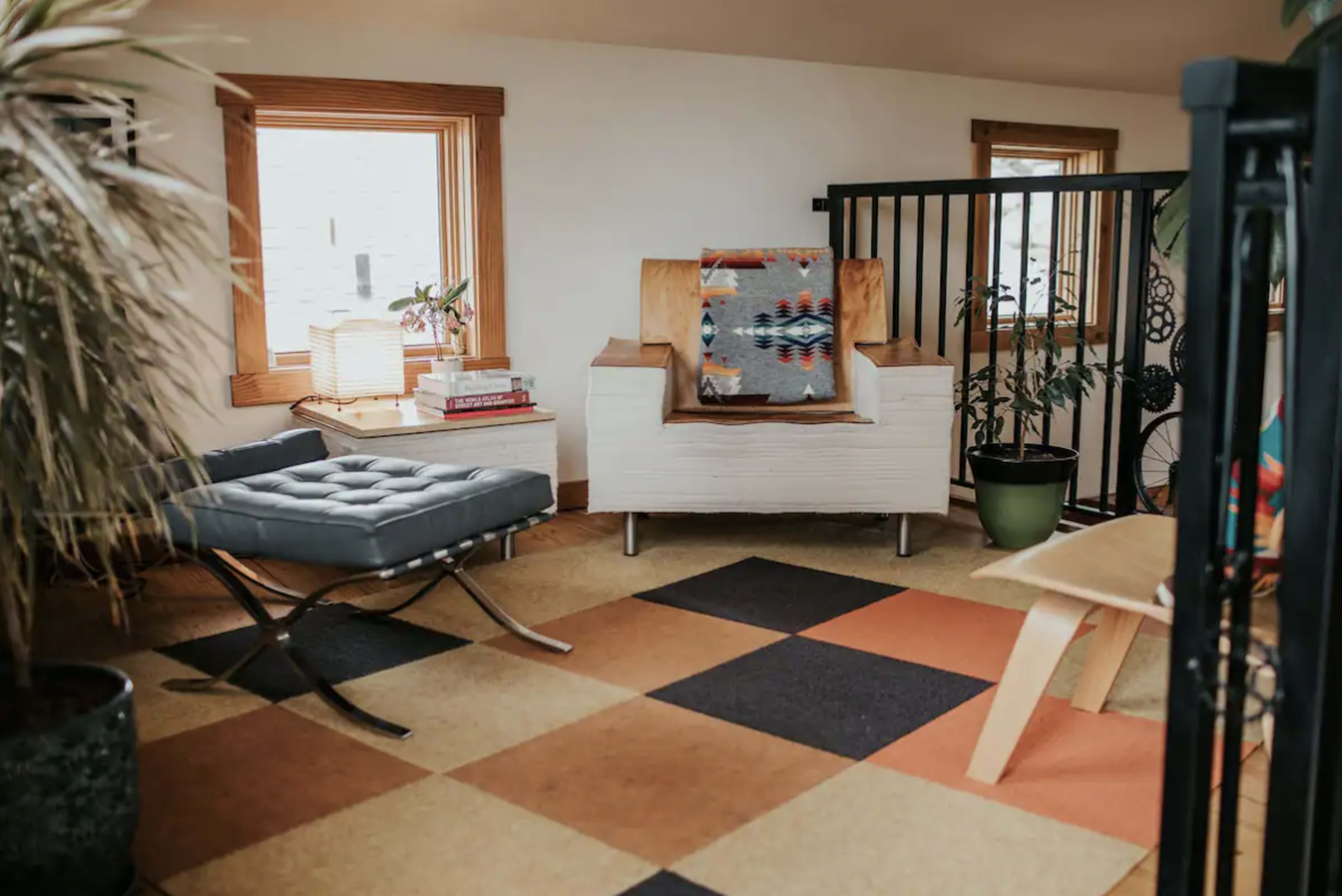 The image shows a cozy room with a modern chair, a small side table, and large windows, featuring a patterned rug and houseplants.