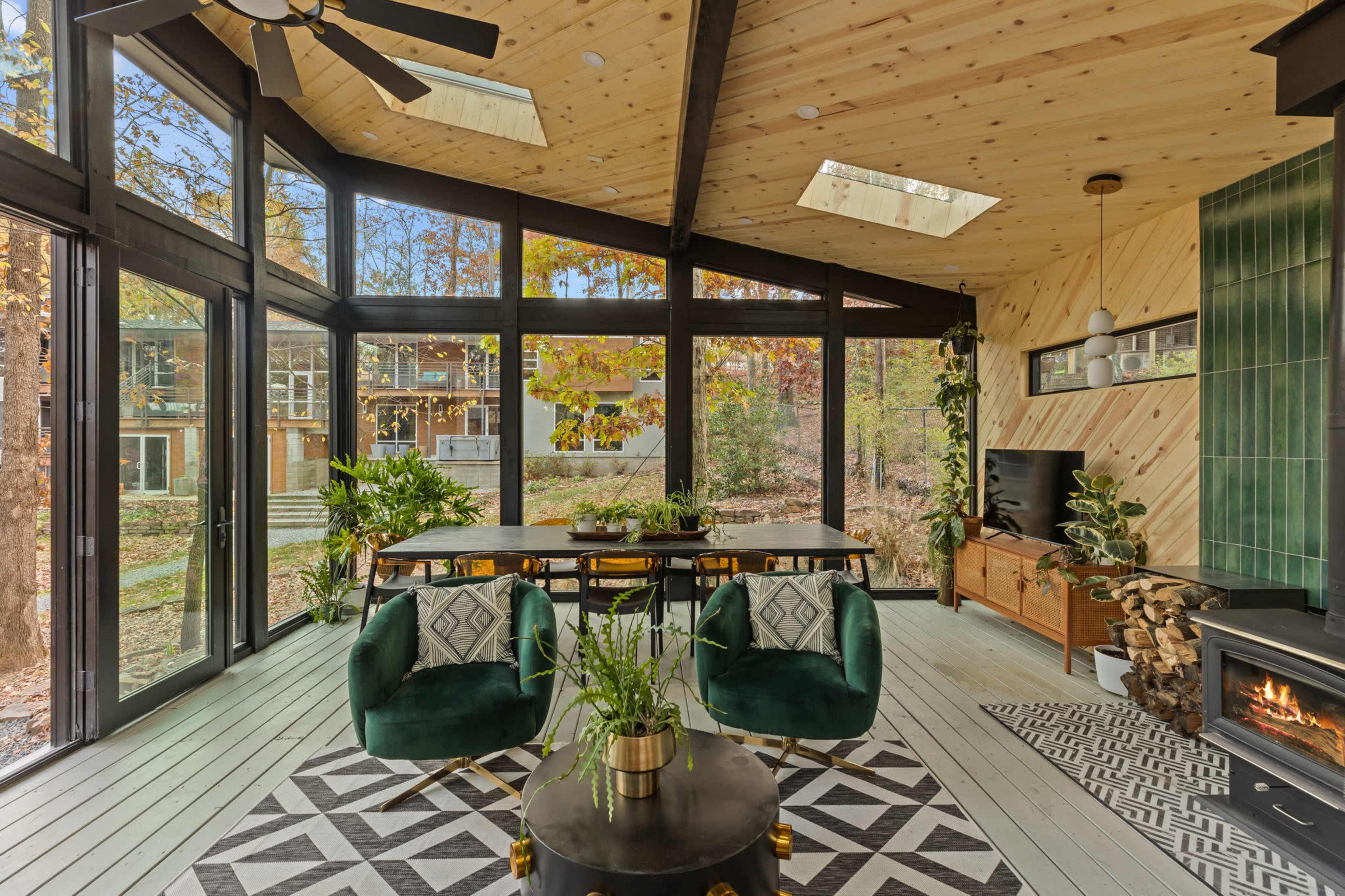A modern interior features large windows, a dining table, green chairs, and a fireplace, surrounded by plants and natural light.