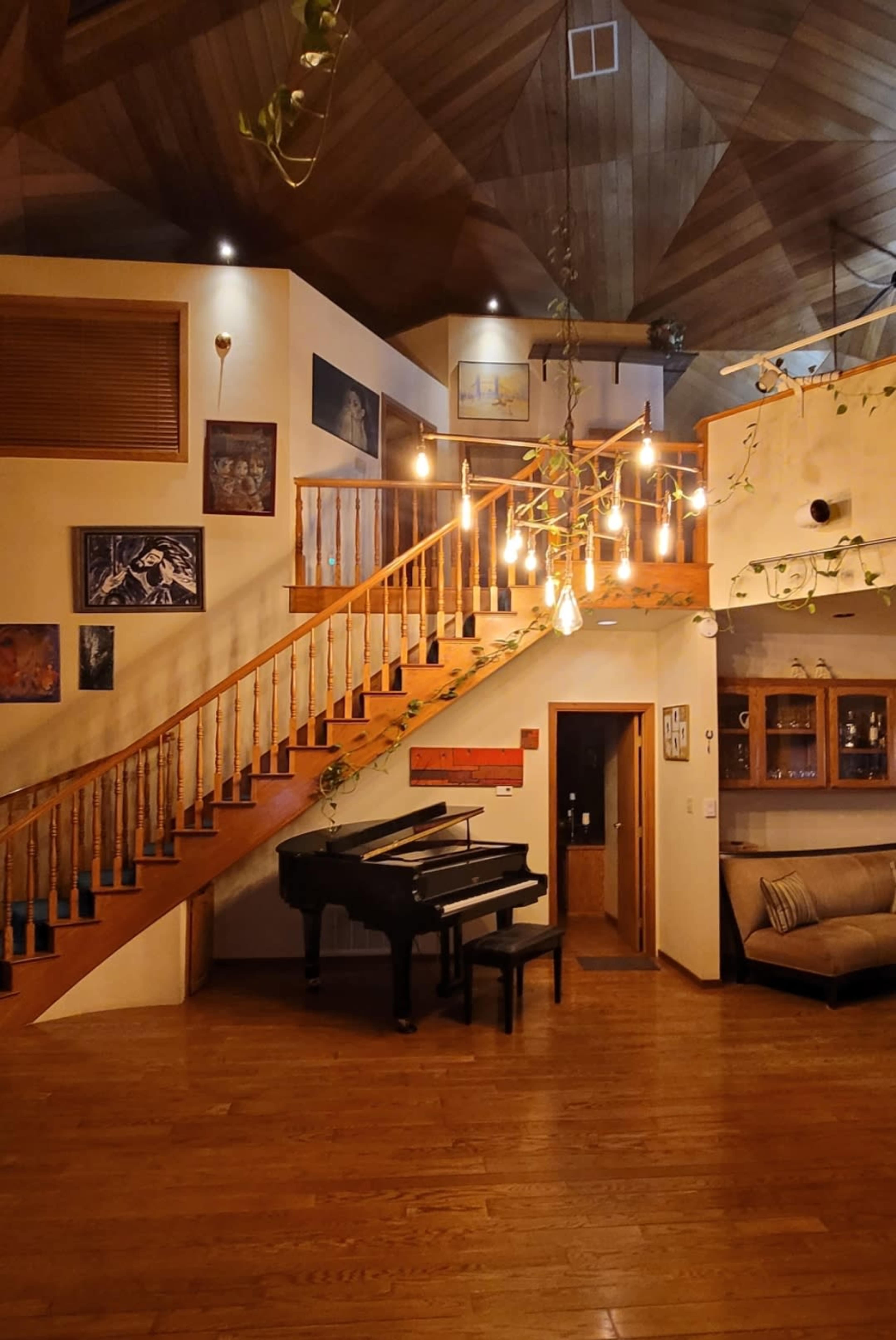 A spacious interior with a grand piano positioned beside a staircase, illuminated by hanging light bulbs and adorned with artwork on the walls.
