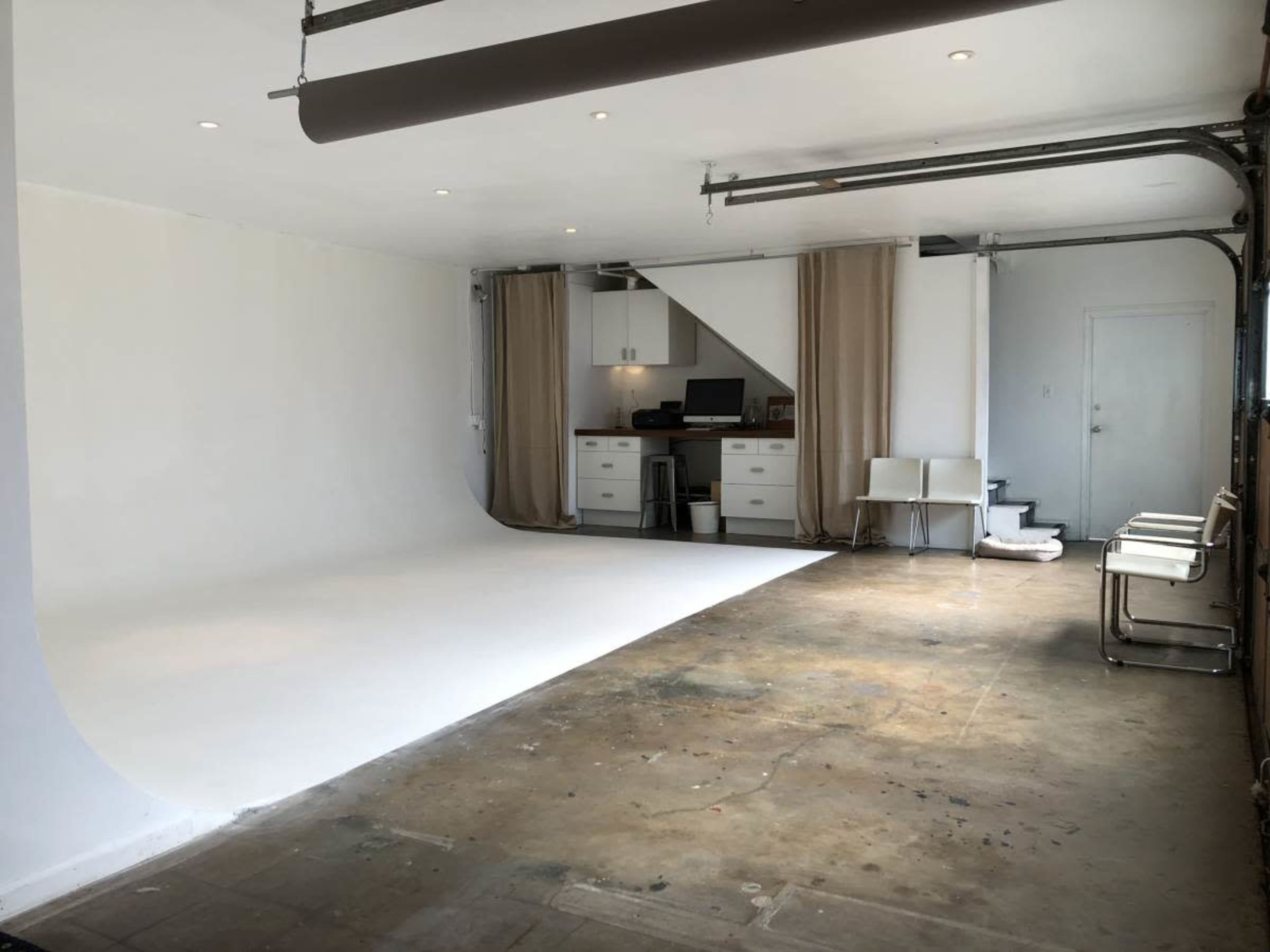 The image shows a spacious photography studio with a seamless white backdrop, minimal furnishings, and a workspace area with a computer and cabinets.