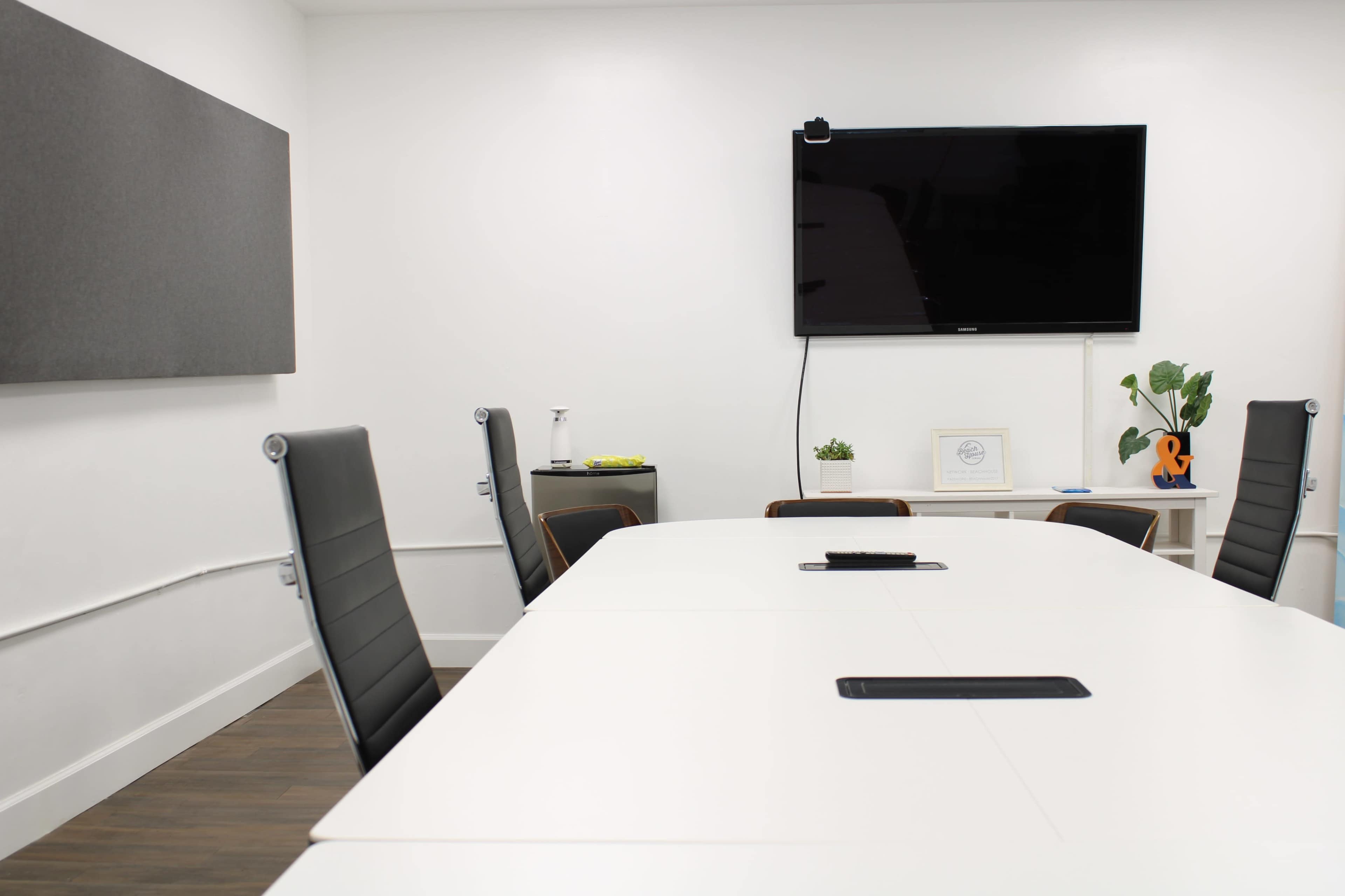 A modern conference room features a long white table, black chairs, a television mounted on the wall, and a small plant on a shelf.