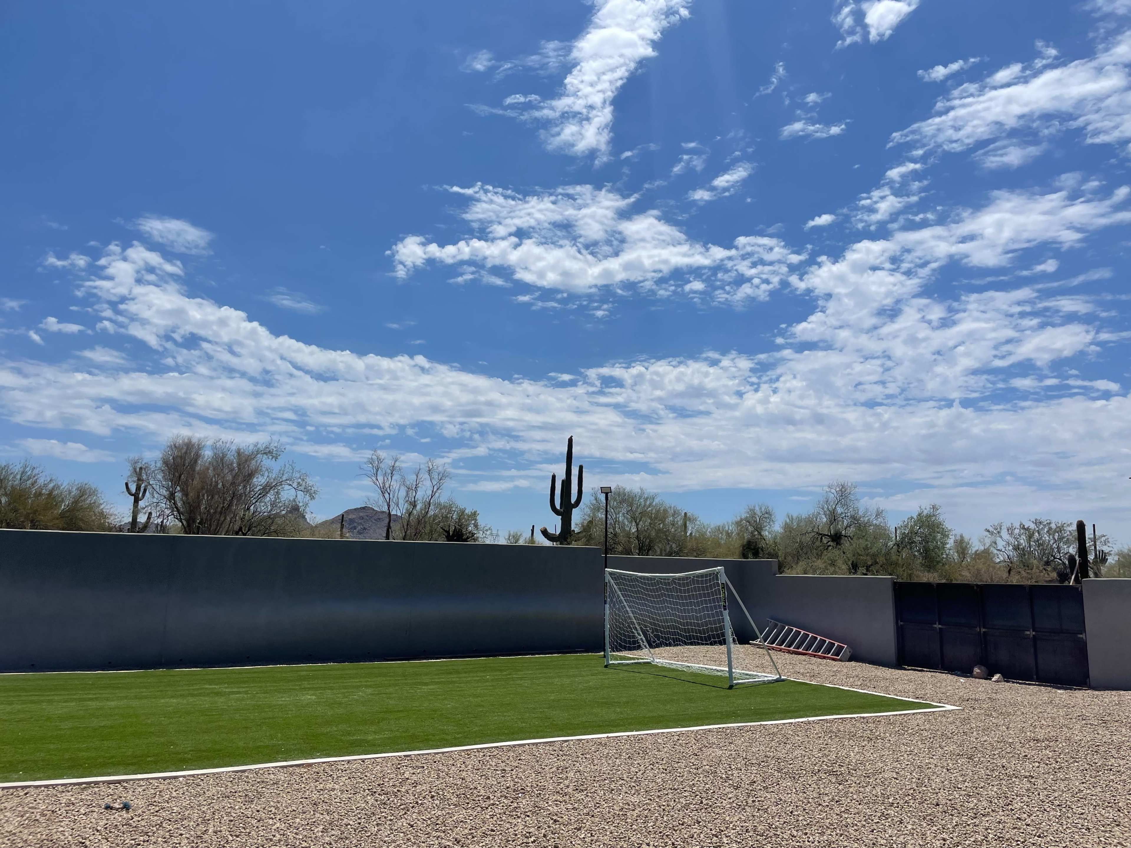 The image shows a small soccer field with a goalpost surrounded by gravel, under a blue sky with scattered clouds and saguaro cacti in the background.