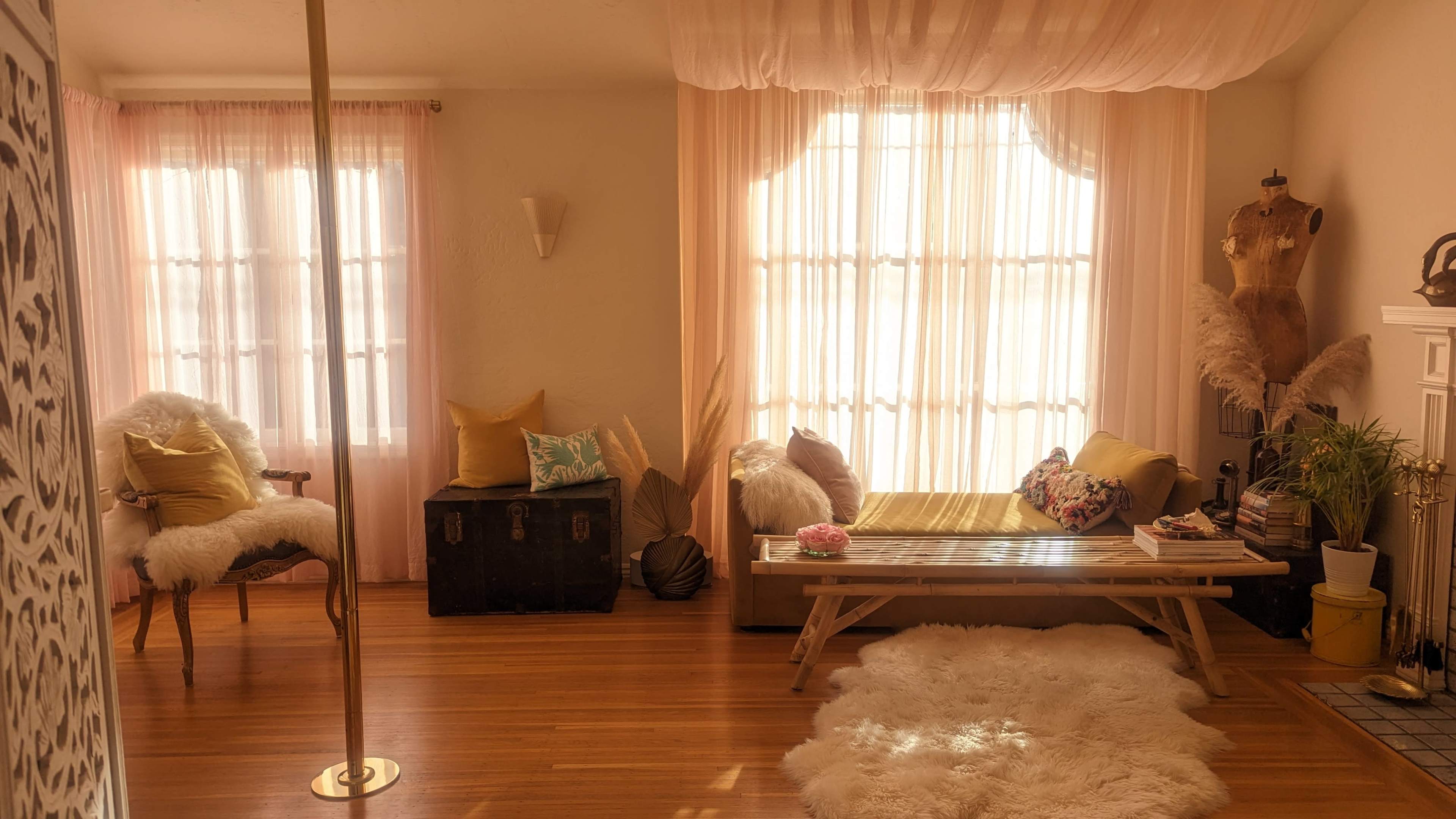 A softly lit room with large windows covered by sheer curtains, a wooden bench, a cozy seating area, and a decorative pole in the corner.