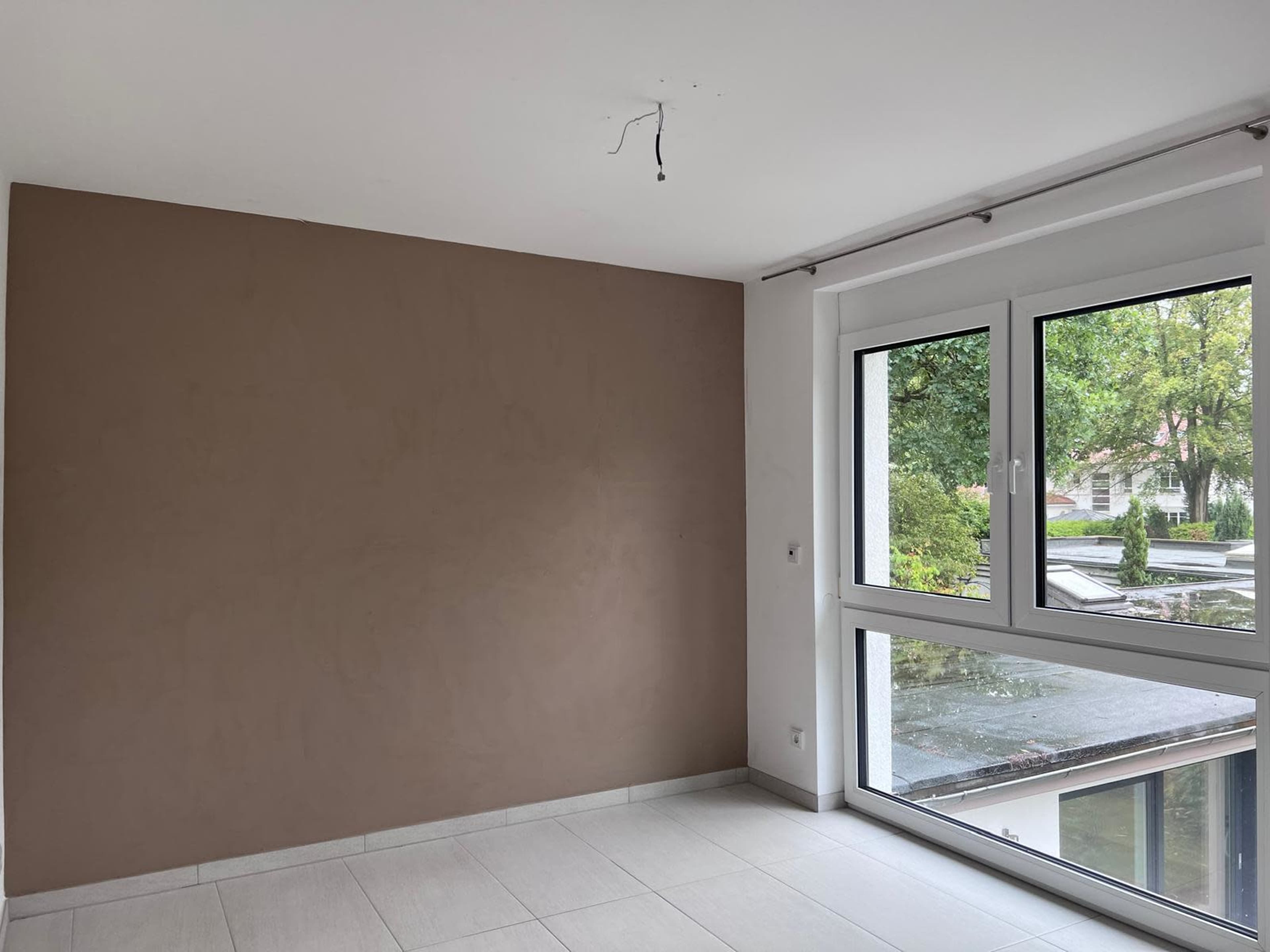 The image shows a bare room with a brown accent wall, large windows, and tiled flooring.