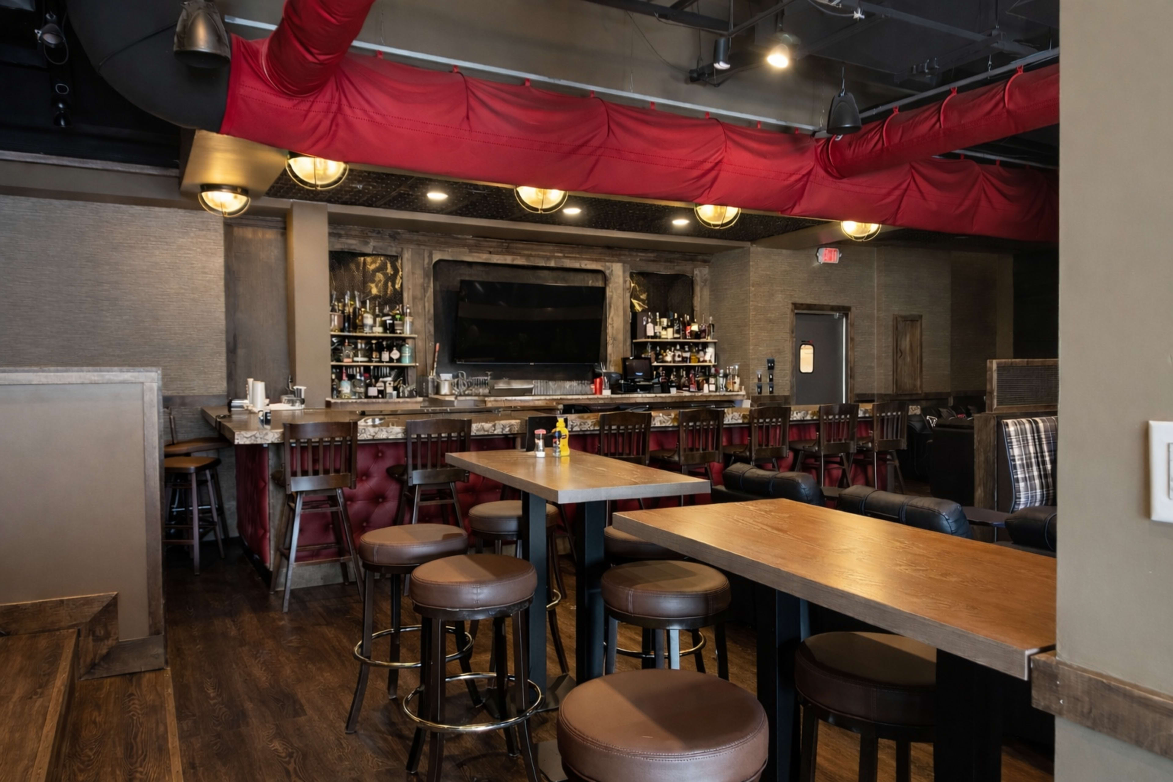 The image shows a modern bar with a long counter, wooden tables, and plush seating against a backdrop of shelves stocked with various bottles.