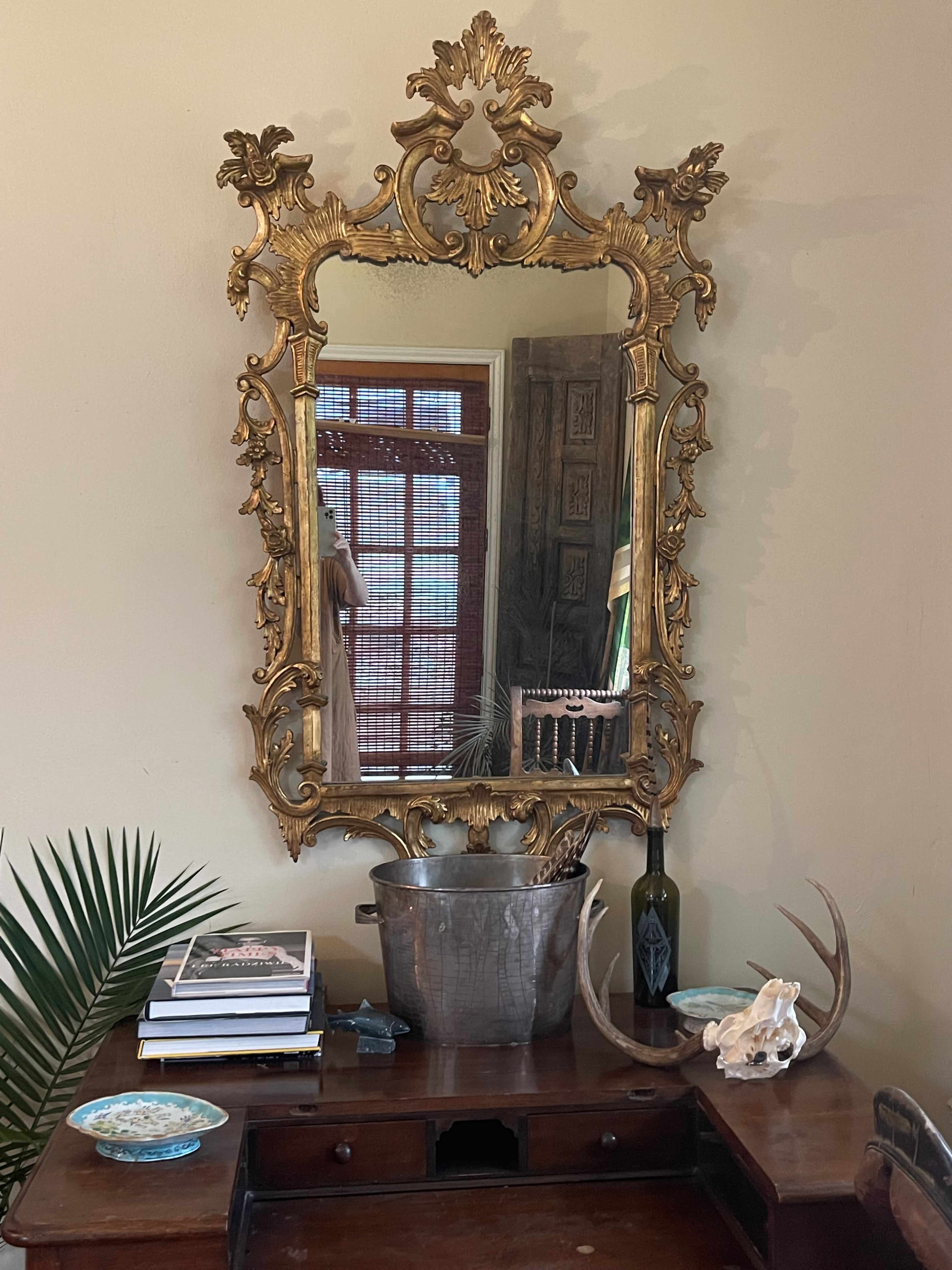 A large ornate gold mirror hangs above a wooden desk, reflecting the room and various objects including a metal bucket, a bottle, and decorative items.