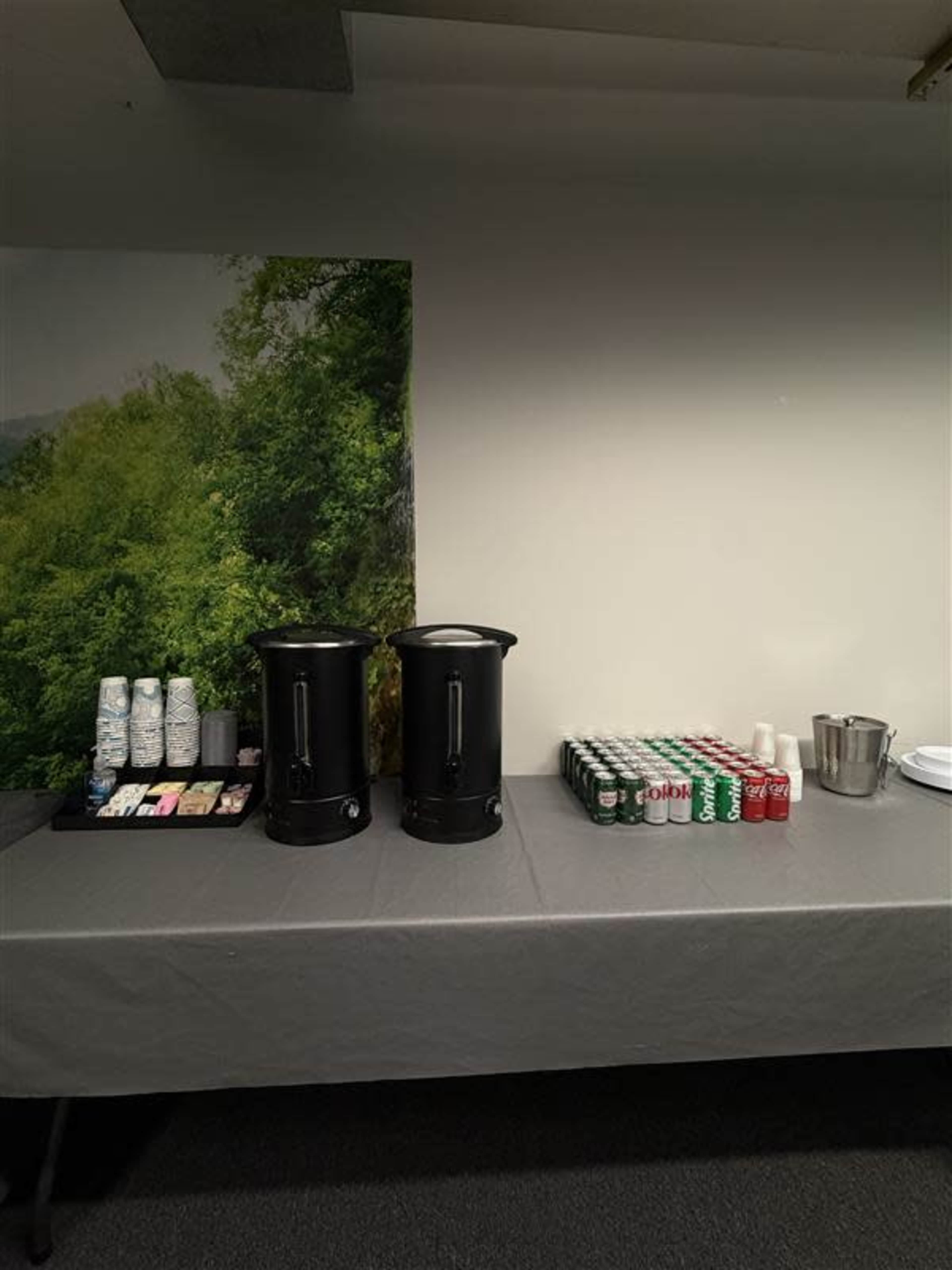 The image shows a table with two black beverage dispensers, a tray of coffee cups and sweeteners, and a row of soda cans.