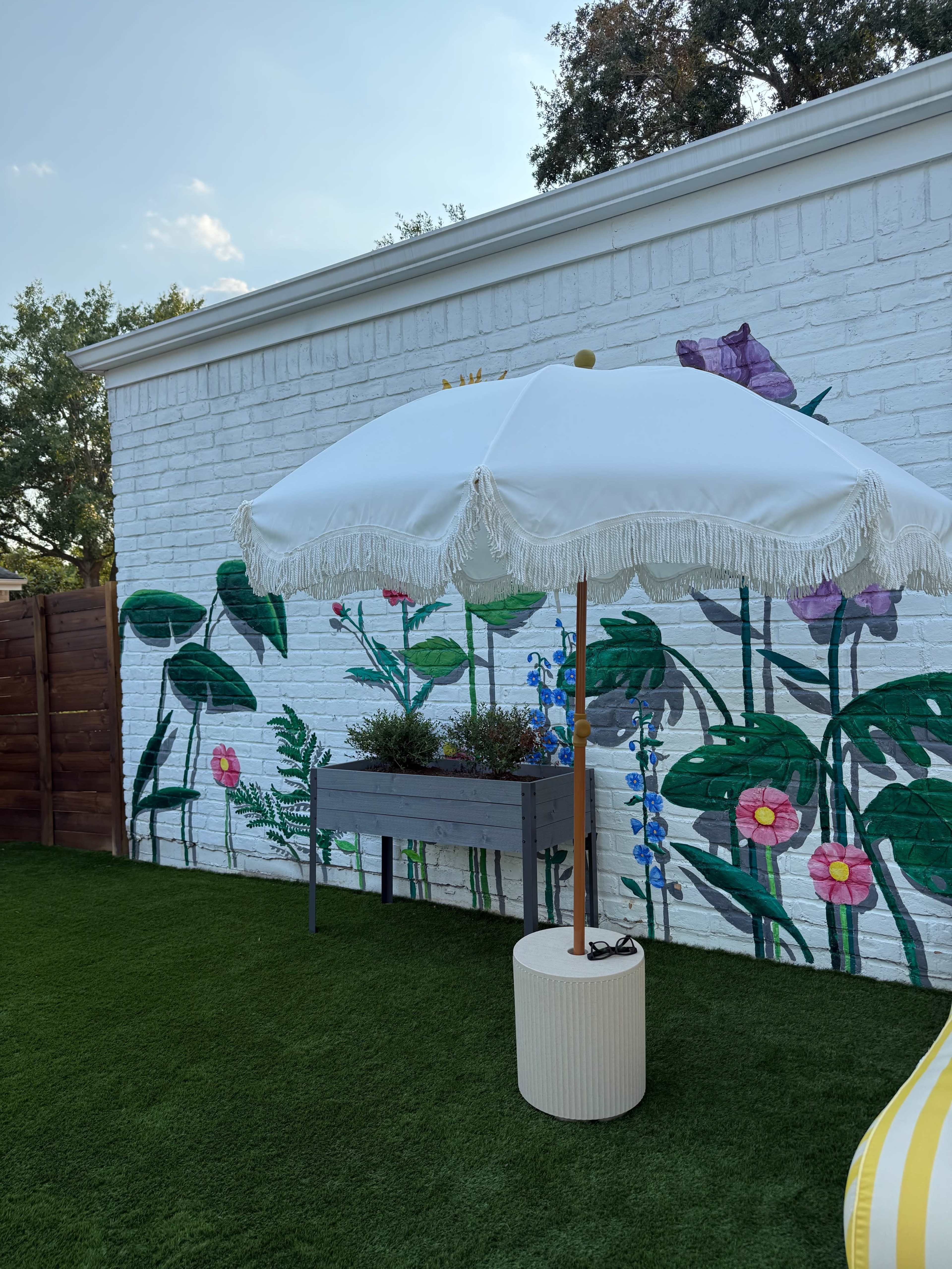 A white umbrella with fringe stands next to a raised planter filled with flowers against a mural of large plants on a white brick wall.