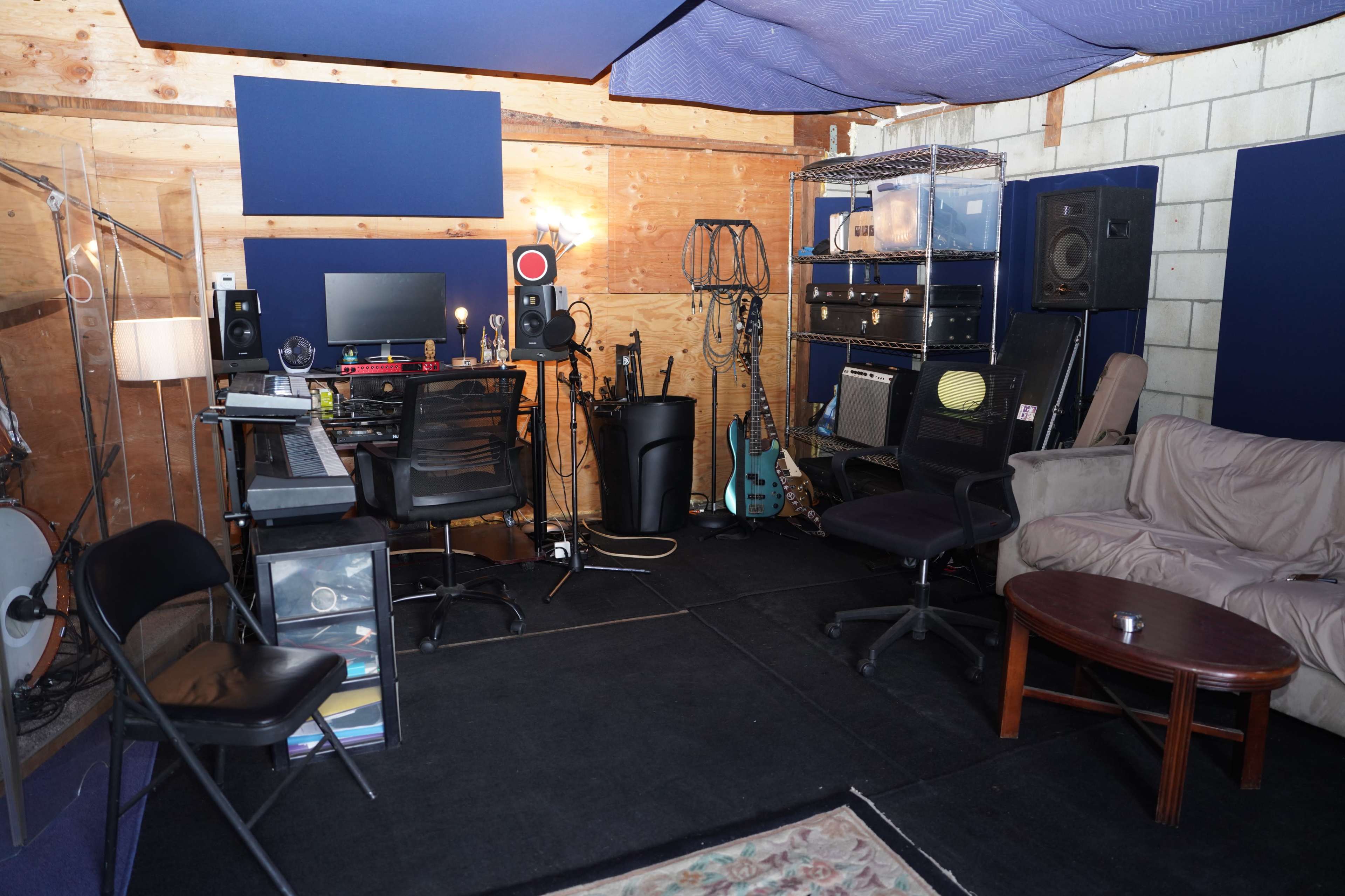 The image shows a small music studio with recording equipment, instruments, and seating arranged around a central workspace.