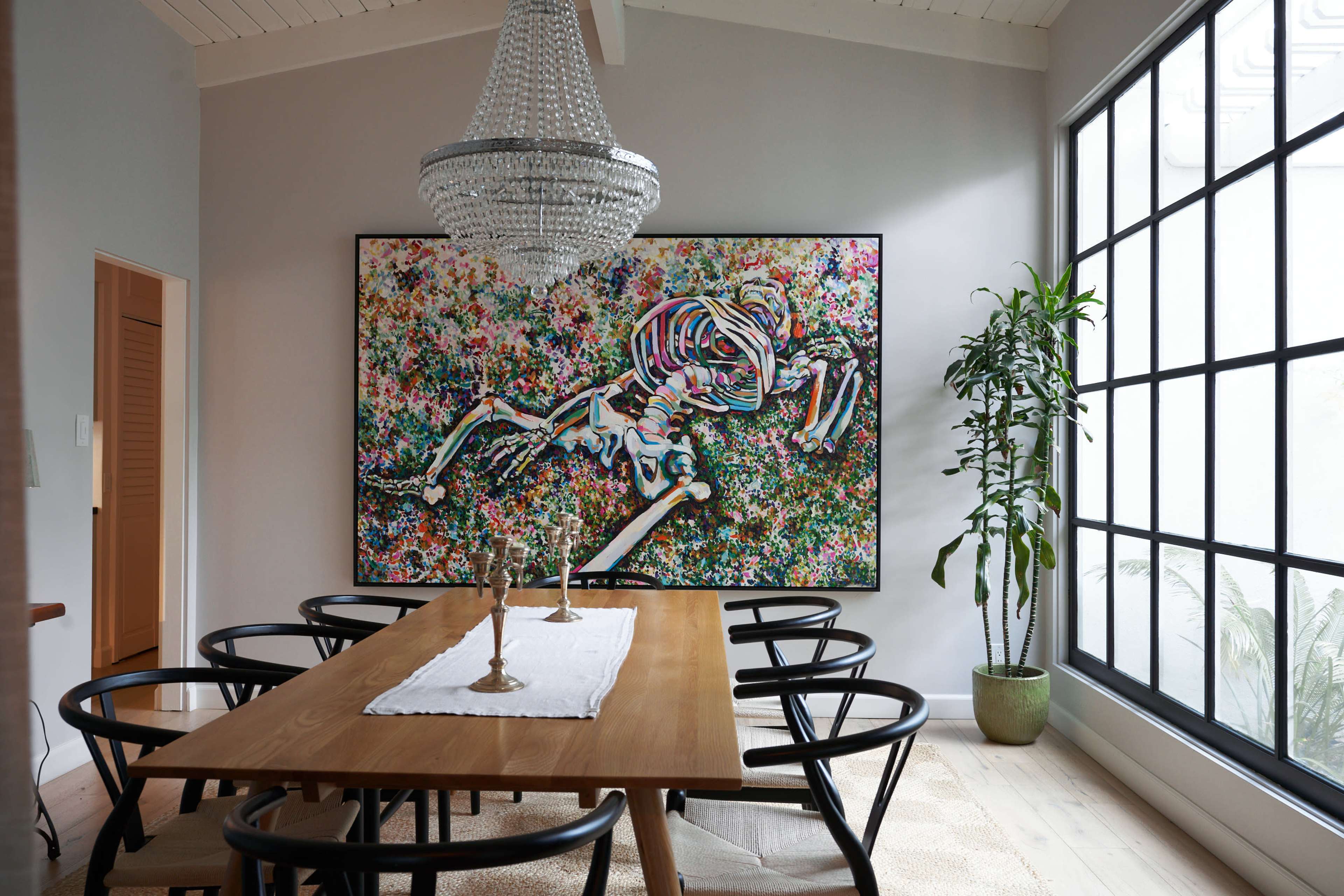 A modern dining room features a large wooden table surrounded by black chairs, with a chandelier above and a colorful artwork of a skeleton mounted on the wall.