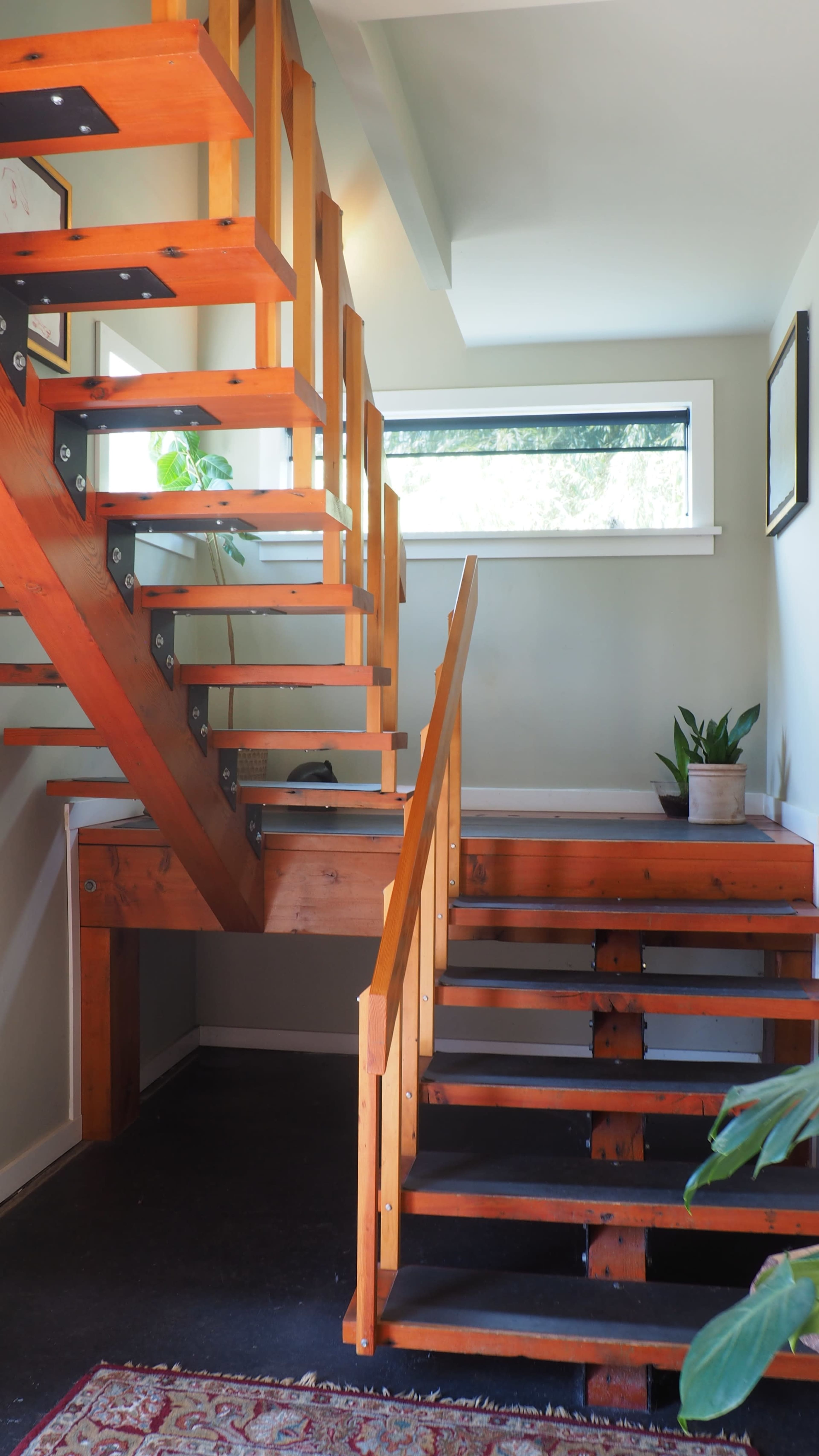 The image shows a modern staircase made of orange wood with metal supports, leading to a landing and a large window above.