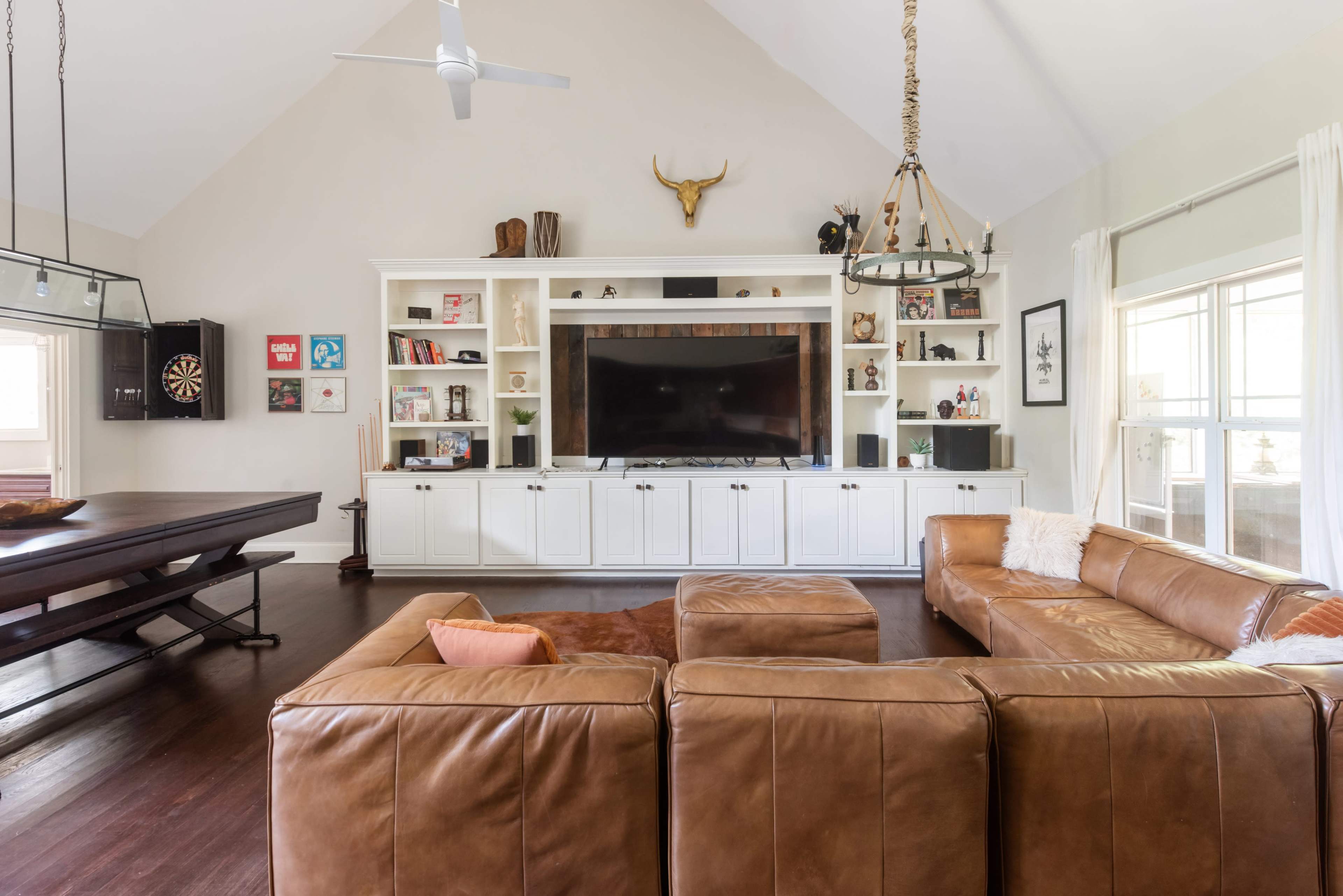 The image shows a spacious living room with a large sectional sofa, a wooden dining table, and a built-in entertainment center featuring a television and decorative items.