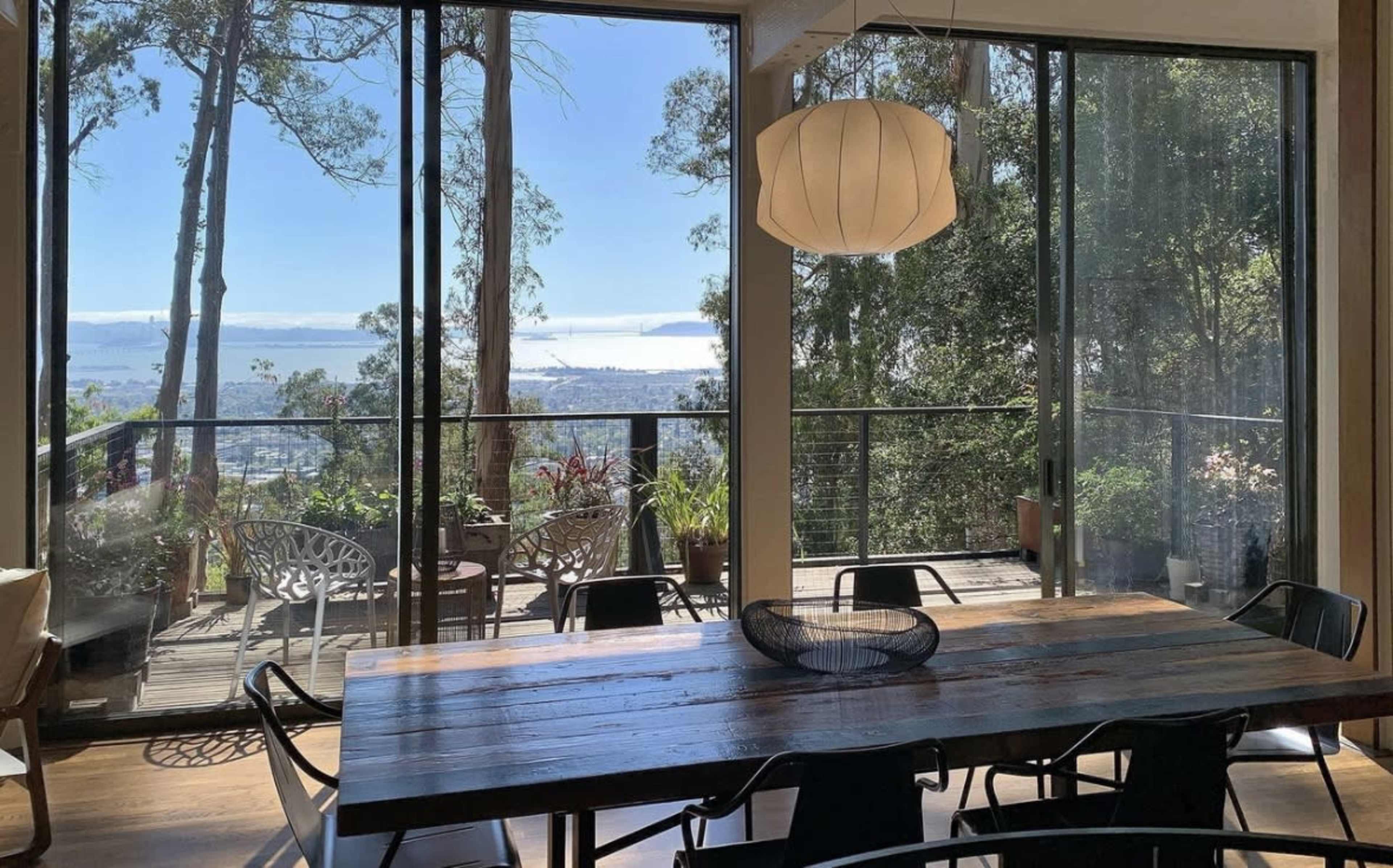 A dining area features a wooden table surrounded by black chairs, with large windows offering a view of trees and a distant body of water.