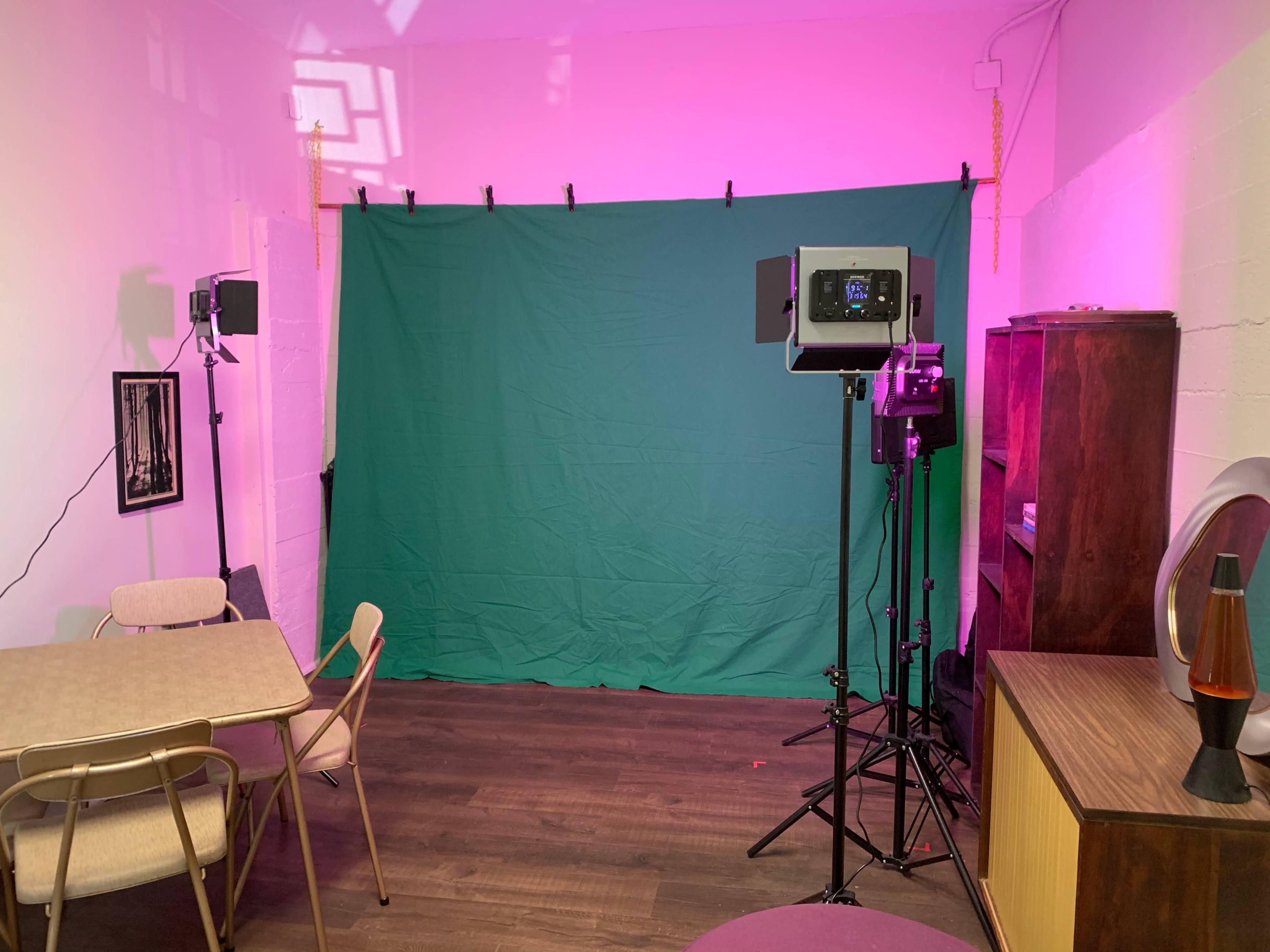 A small room is set up with a green backdrop, lighting equipment, a table, and a wooden cabinet.