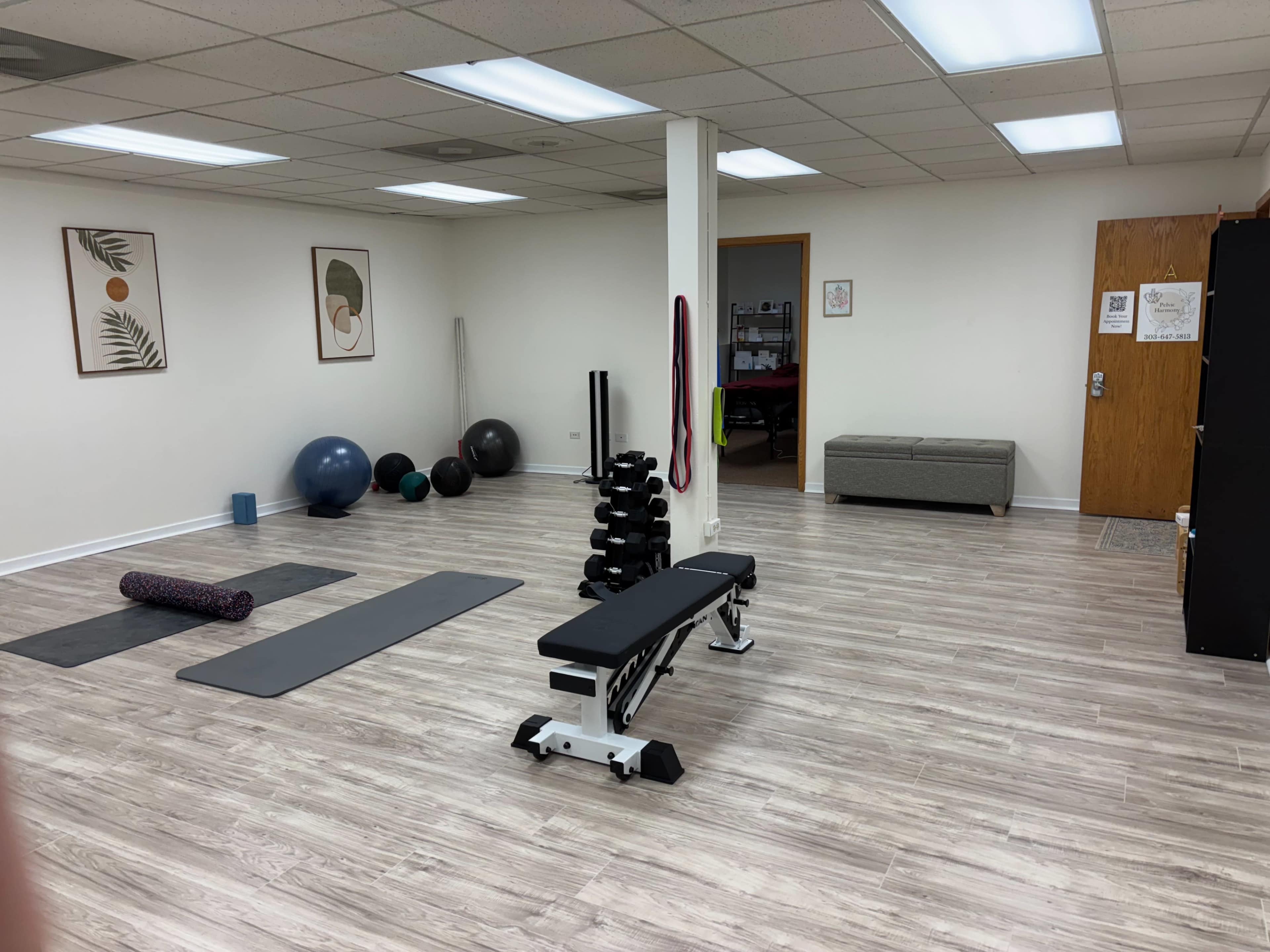 The image shows a spacious fitness room with exercise mats, weights, stability balls, and a bench, illuminated by overhead lights.
