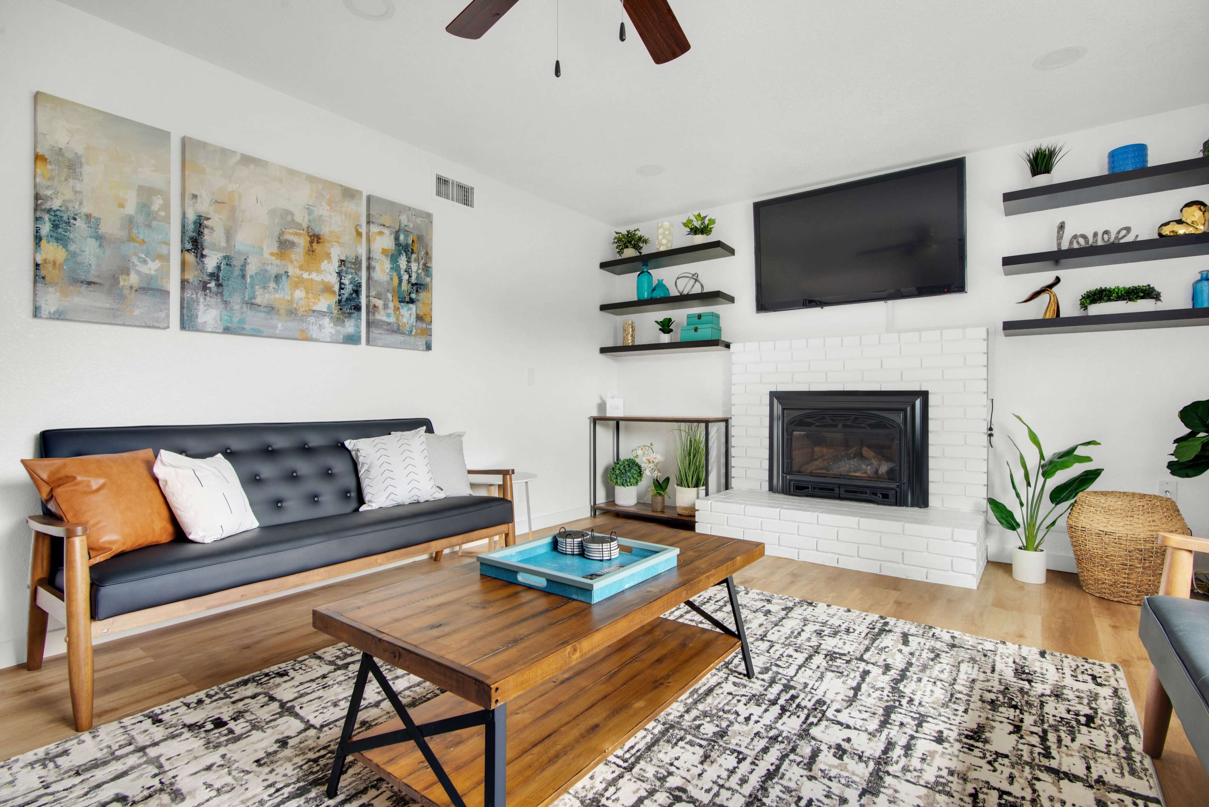 A modern living room features a black sofa, a wooden coffee table, and a white brick fireplace, with abstract artwork on the walls and decorative plants throughout.