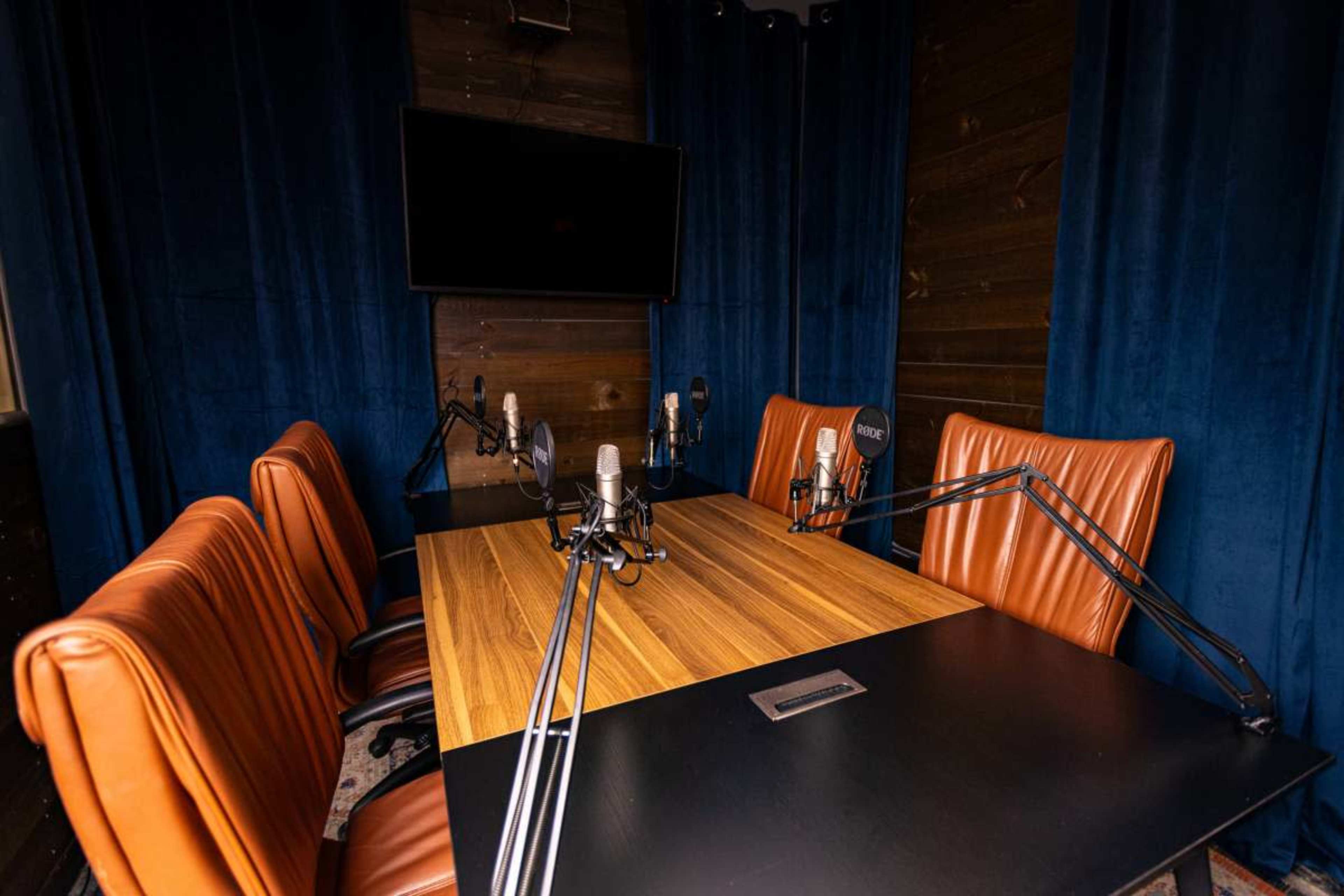 The image shows a soundproof recording studio with a wooden table, four leather chairs, and multiple microphones mounted on adjustable arms.