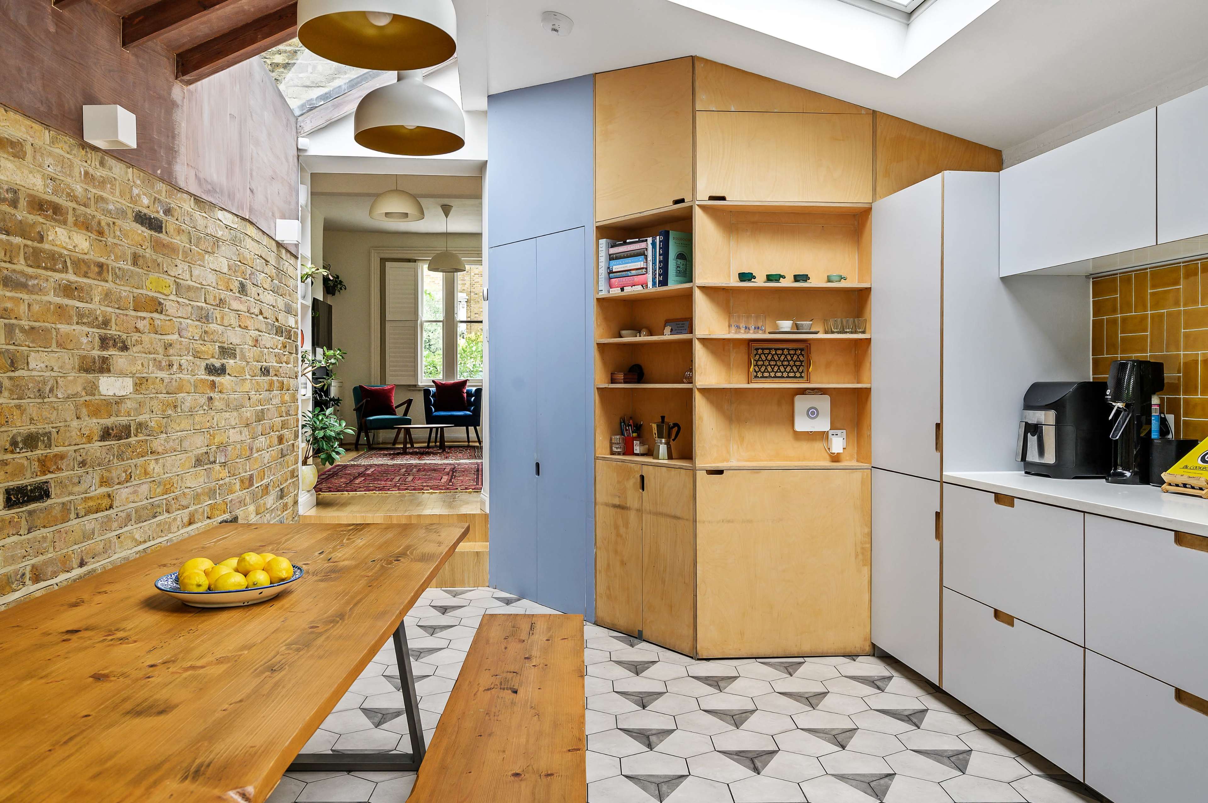 The image shows a modern kitchen with wooden and white cabinetry, a patterned tiled floor, and a wooden dining table with a bowl of lemons.