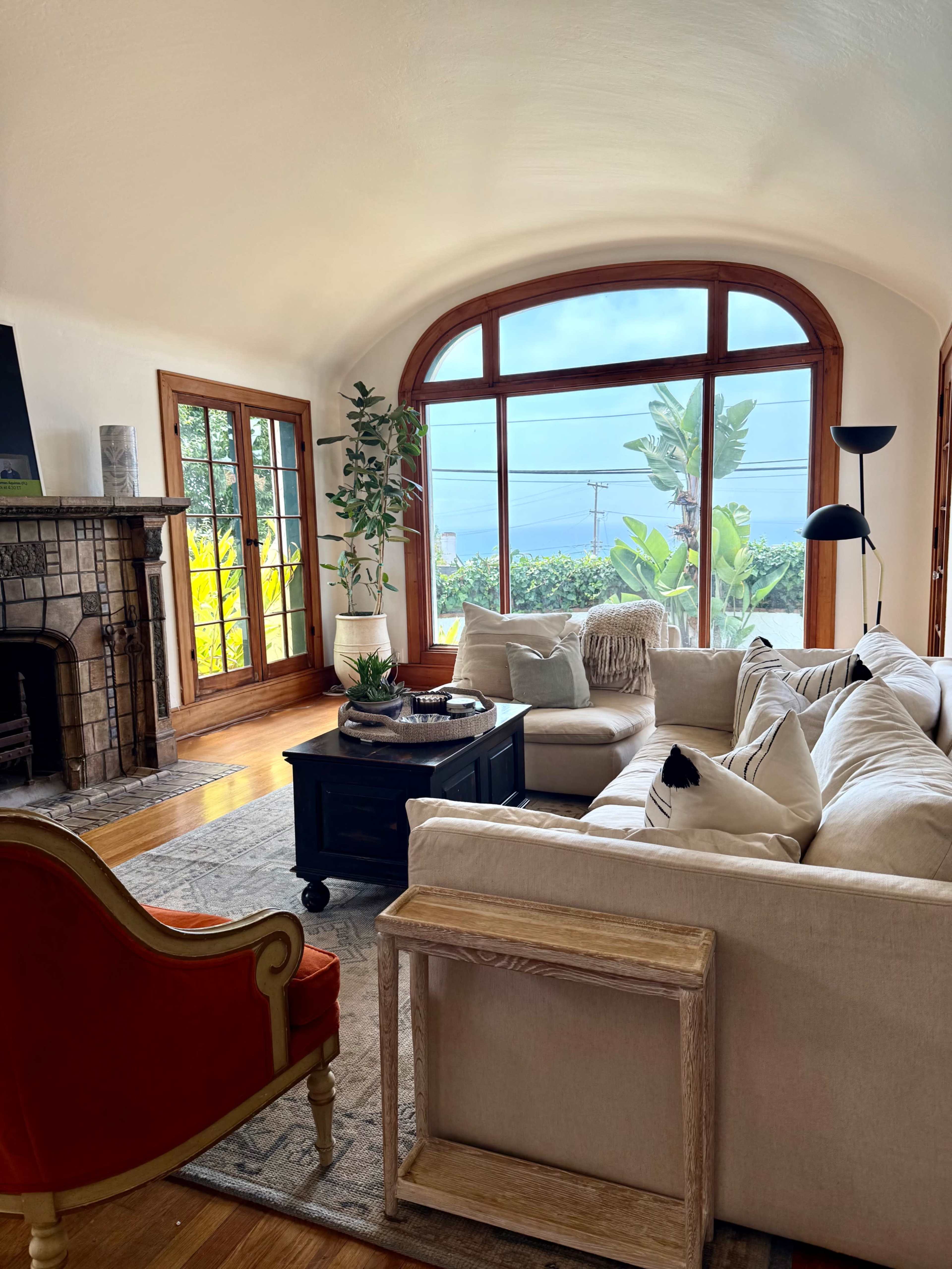 A cozy living room features a large sectional sofa, an armchair, a coffee table, and a fireplace, with large windows offering a view of the outdoors.
