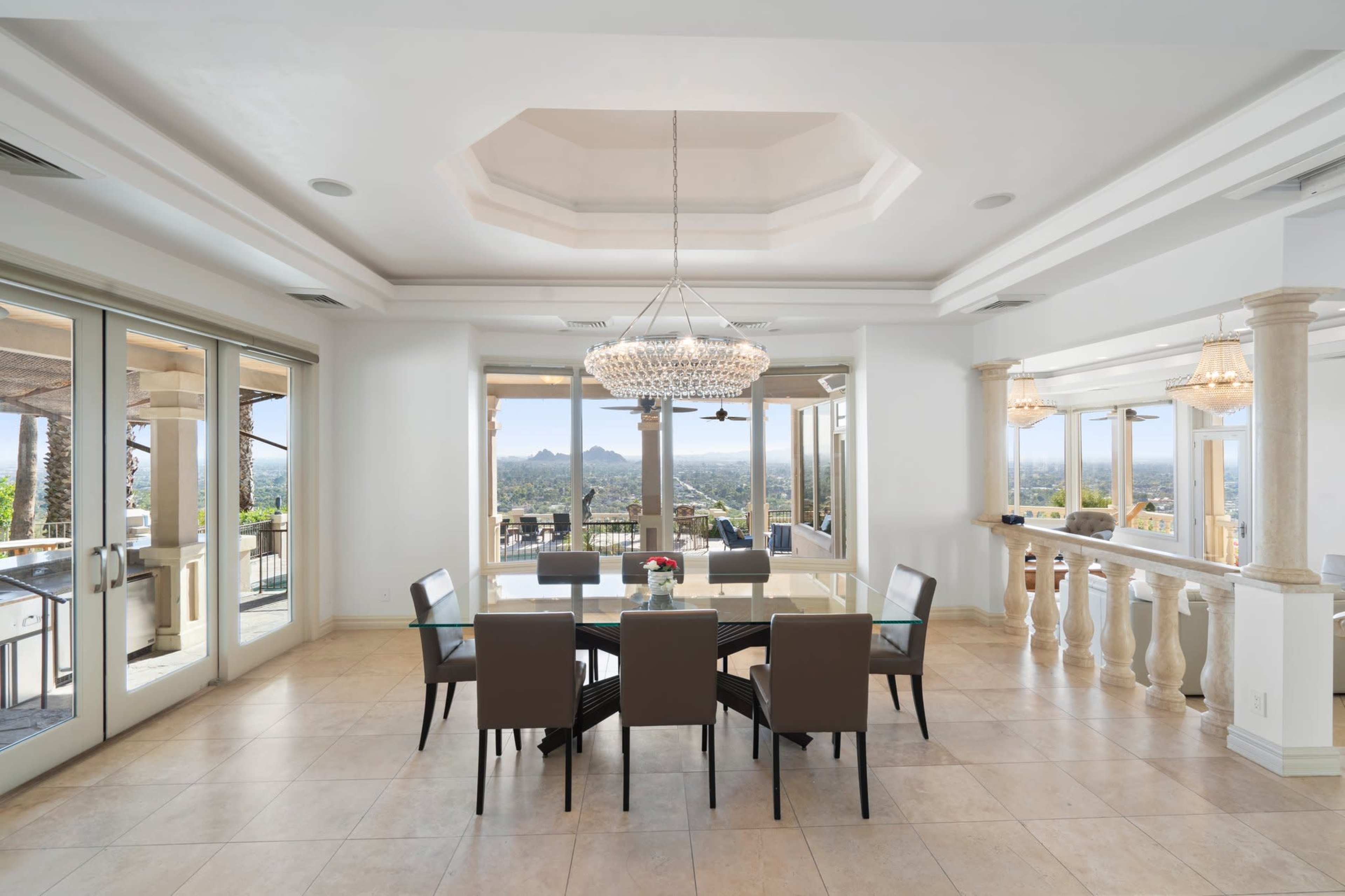 The image shows a spacious dining area featuring a large table surrounded by six chairs, a chandelier hanging from the ceiling, and large windows that offer a panoramic view of the landscape outside.
