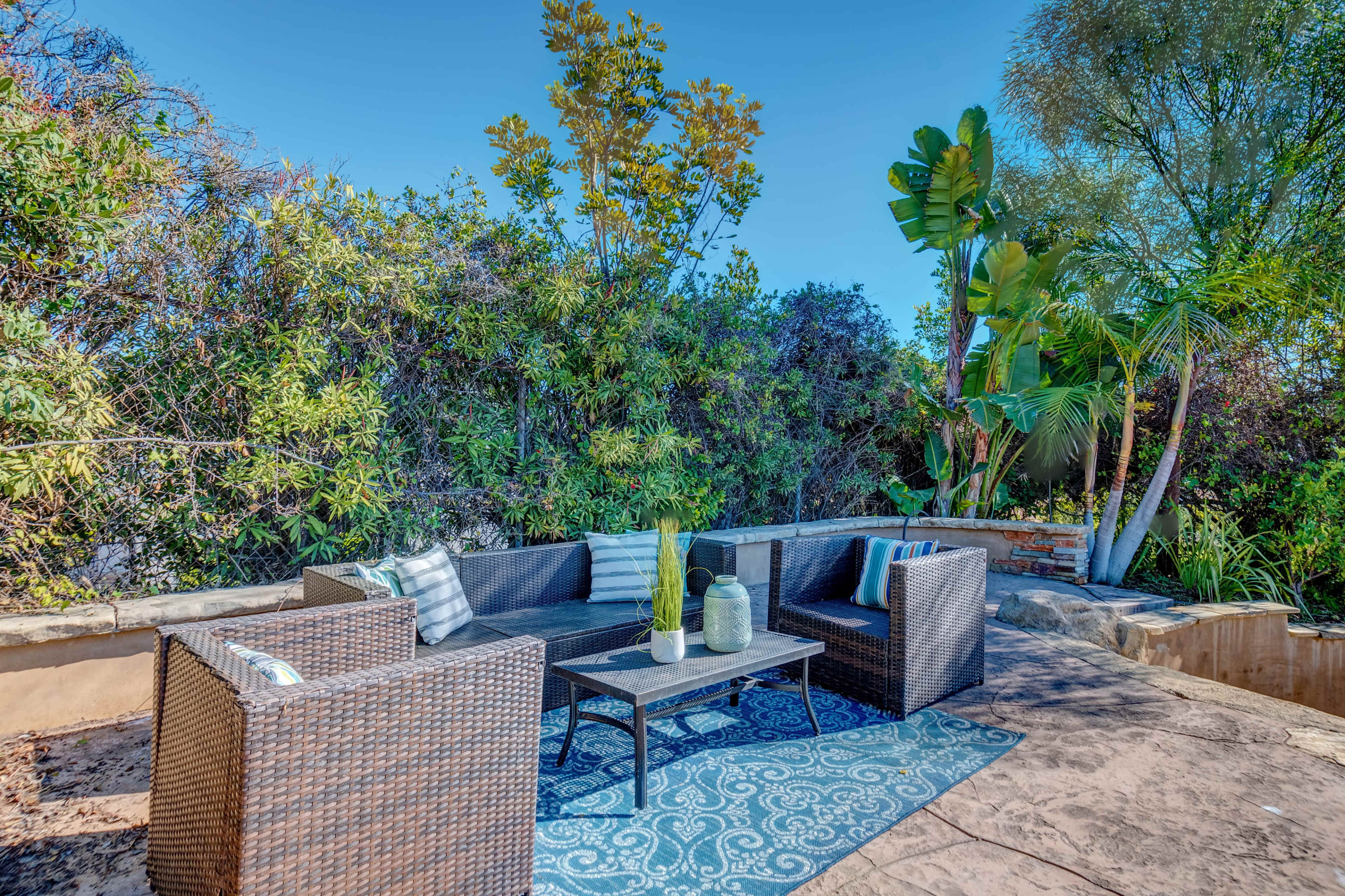 A cozy outdoor seating area features a black wicker sofa set arranged around a coffee table on a patterned rug, surrounded by lush greenery.