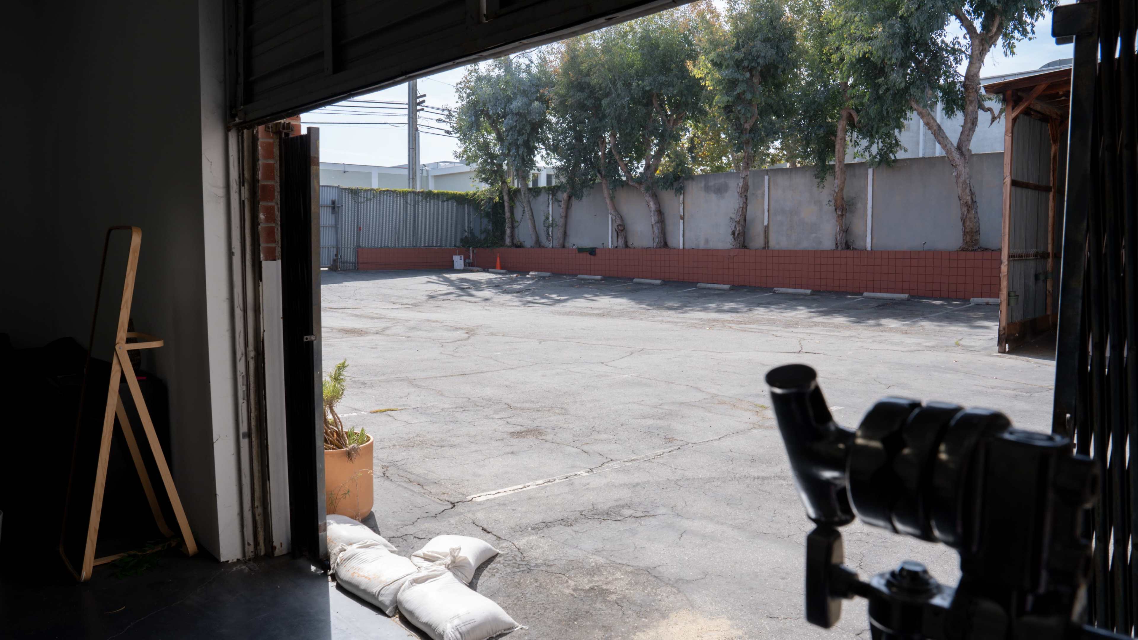 The image shows an open garage door overlooking a vacant, paved parking lot bordered by trees and a wall.