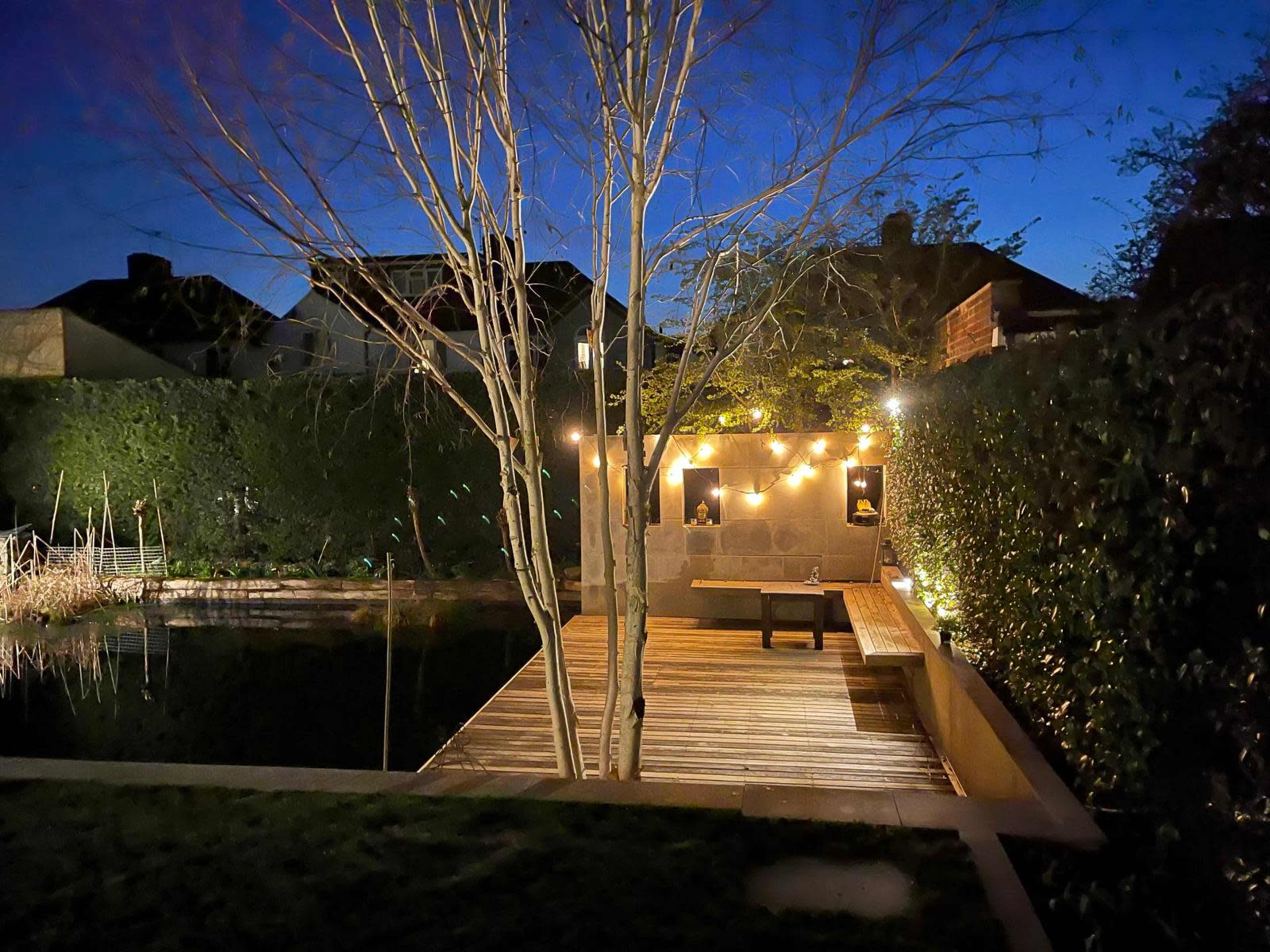 A wooden deck by a pond, illuminated by string lights, surrounded by trees and shrubs at dusk.