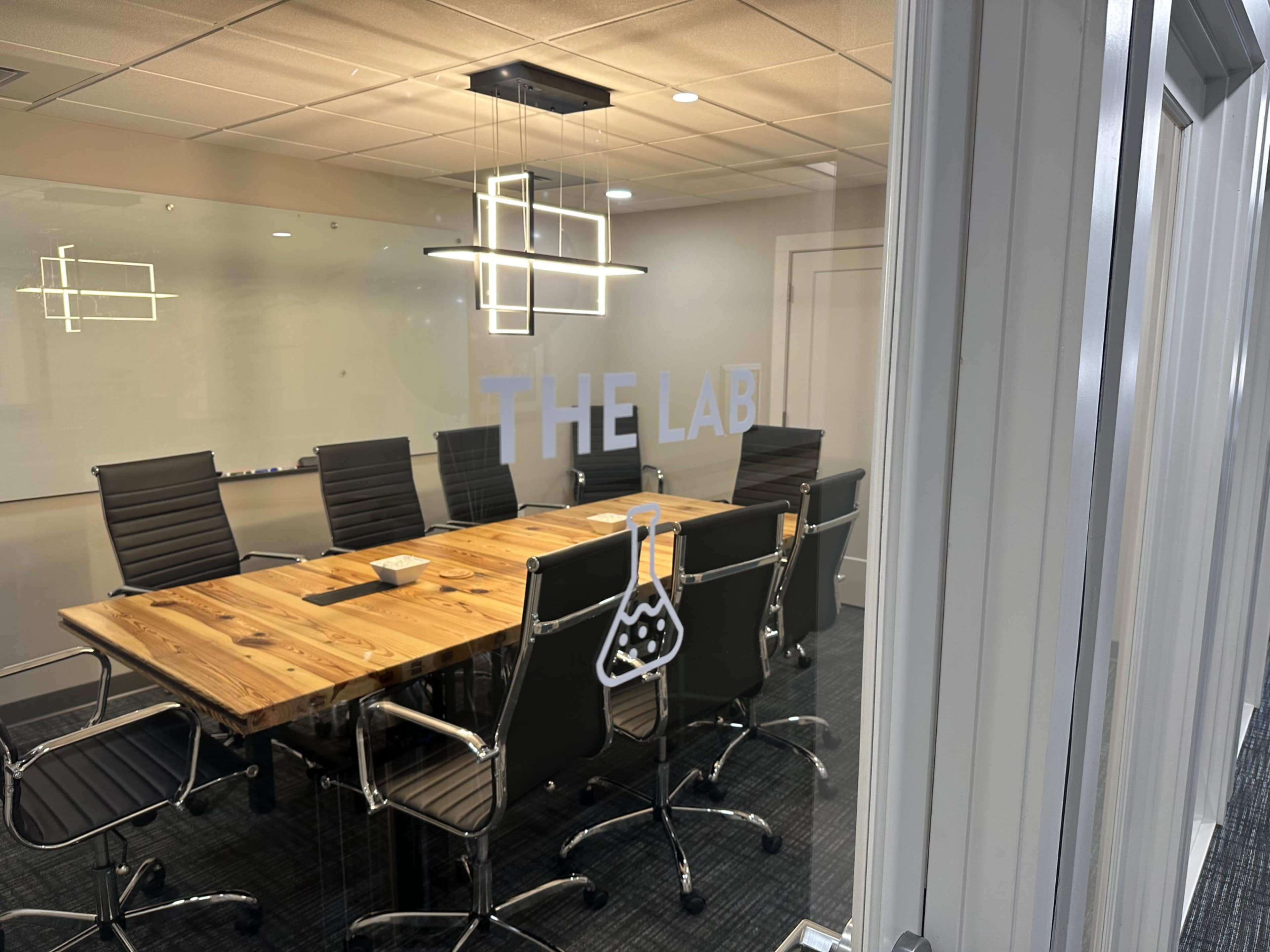 A conference room with a large wooden table surrounded by black leather chairs is visible through glass doors, featuring the words "THE LAB" and a lab flask logo on the glass.