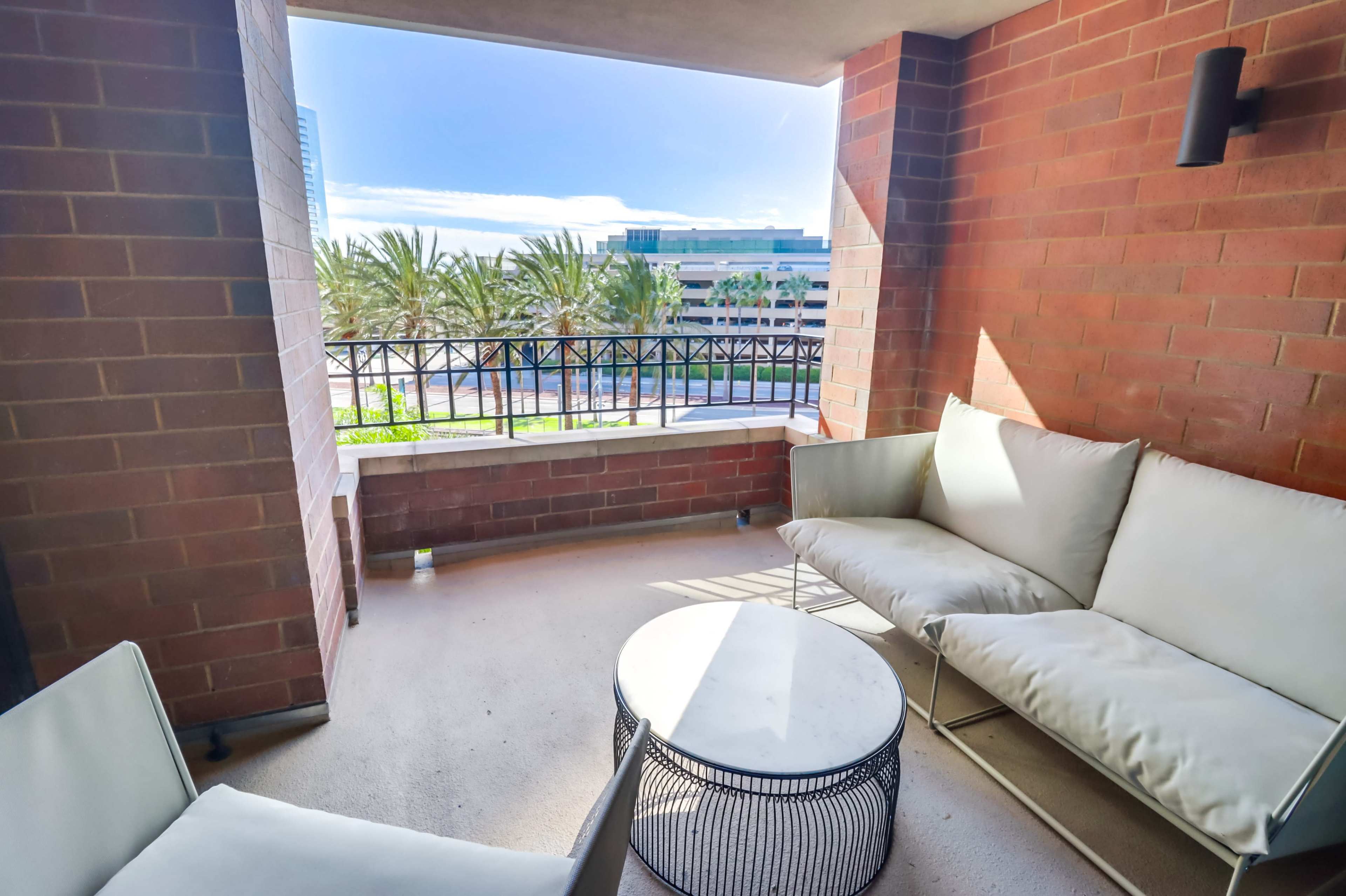 A balcony features a white sofa set and a circular table, overlooking palm trees and a cityscape.