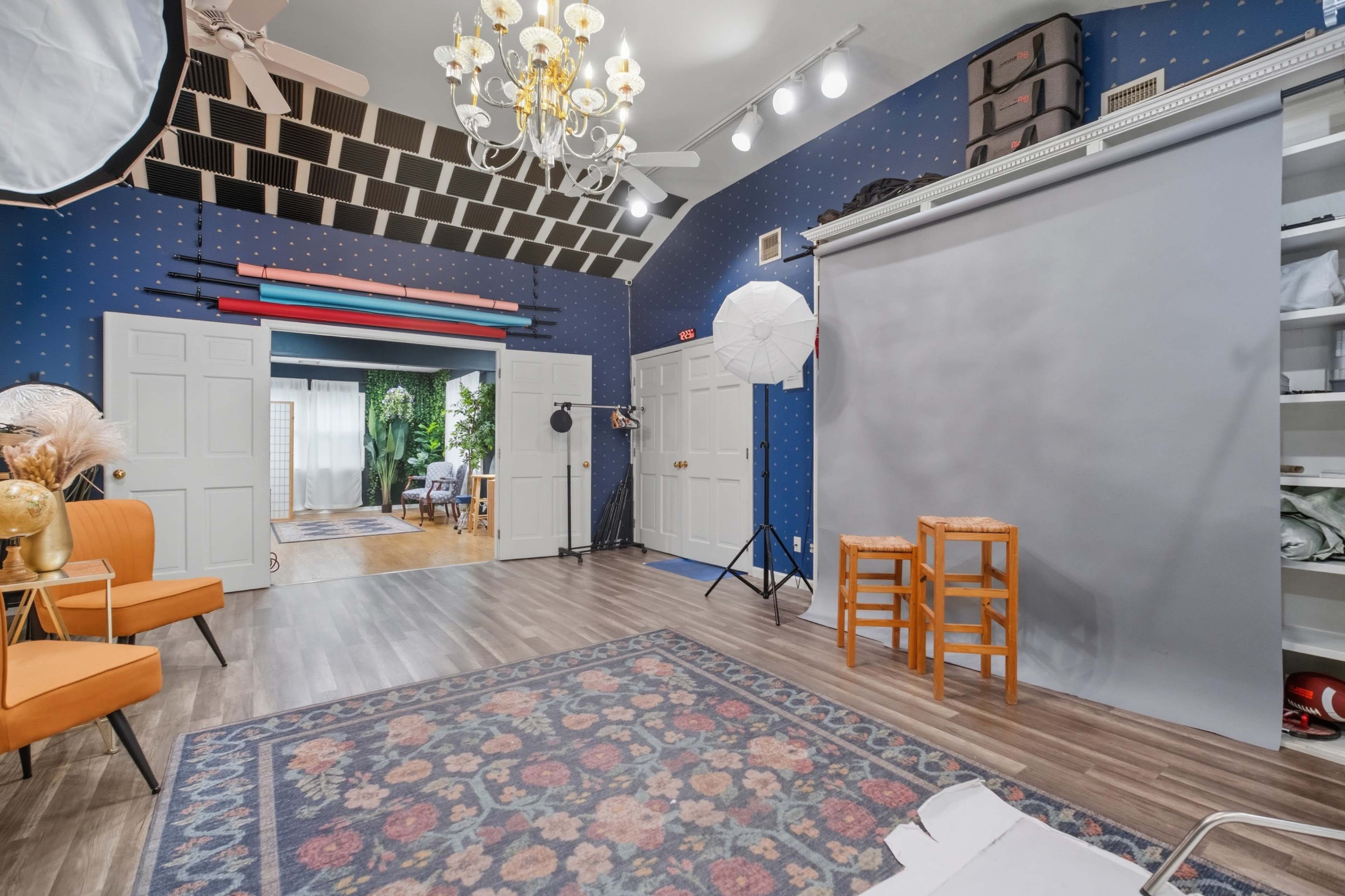 The image shows a well-lit studio with a gray backdrop, various lighting equipment, a patterned rug, and two orange chairs.