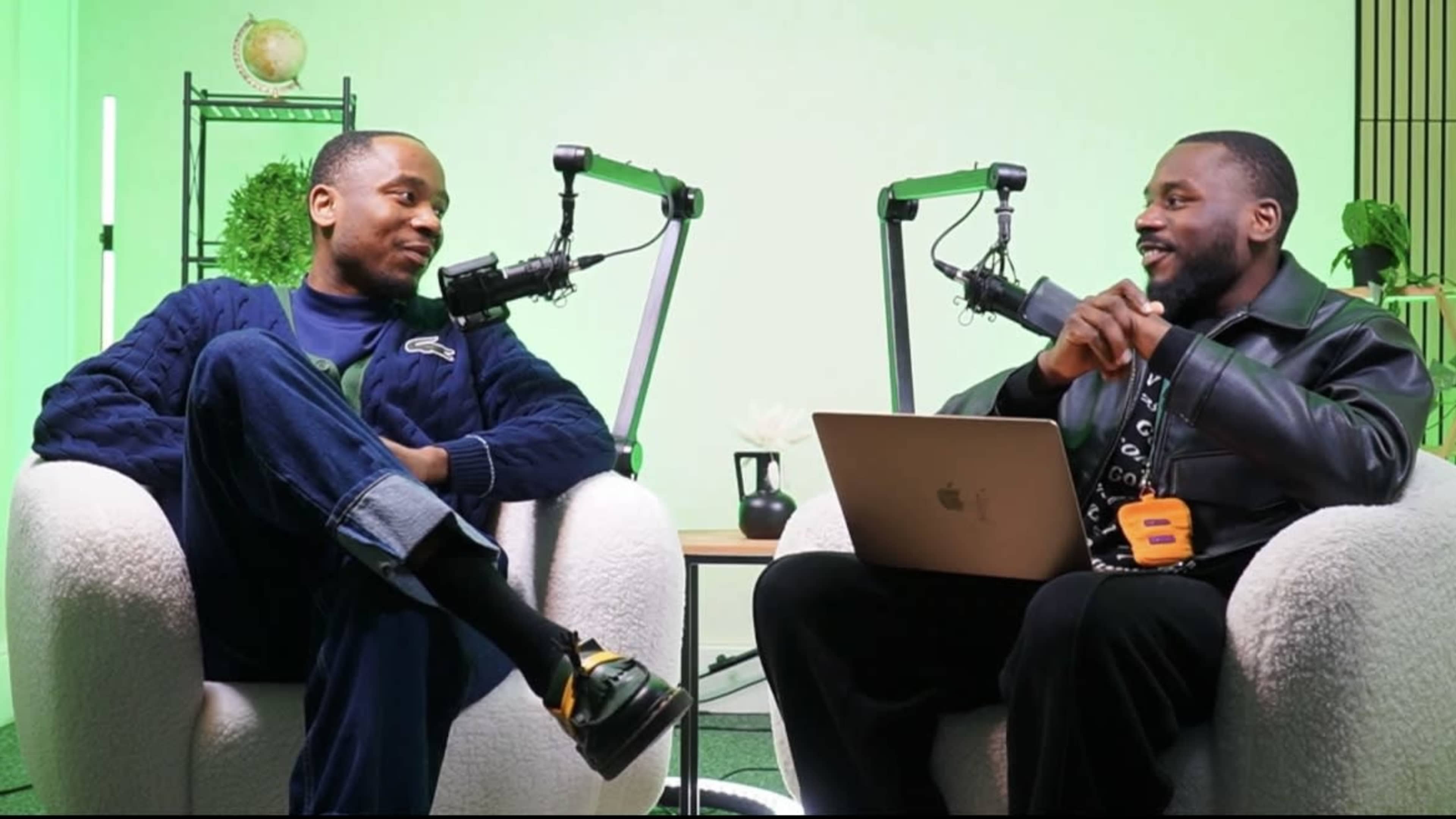 Two men are seated in a modern studio setting with green lighting, discussing while using microphones and a laptop placed on a table between them.