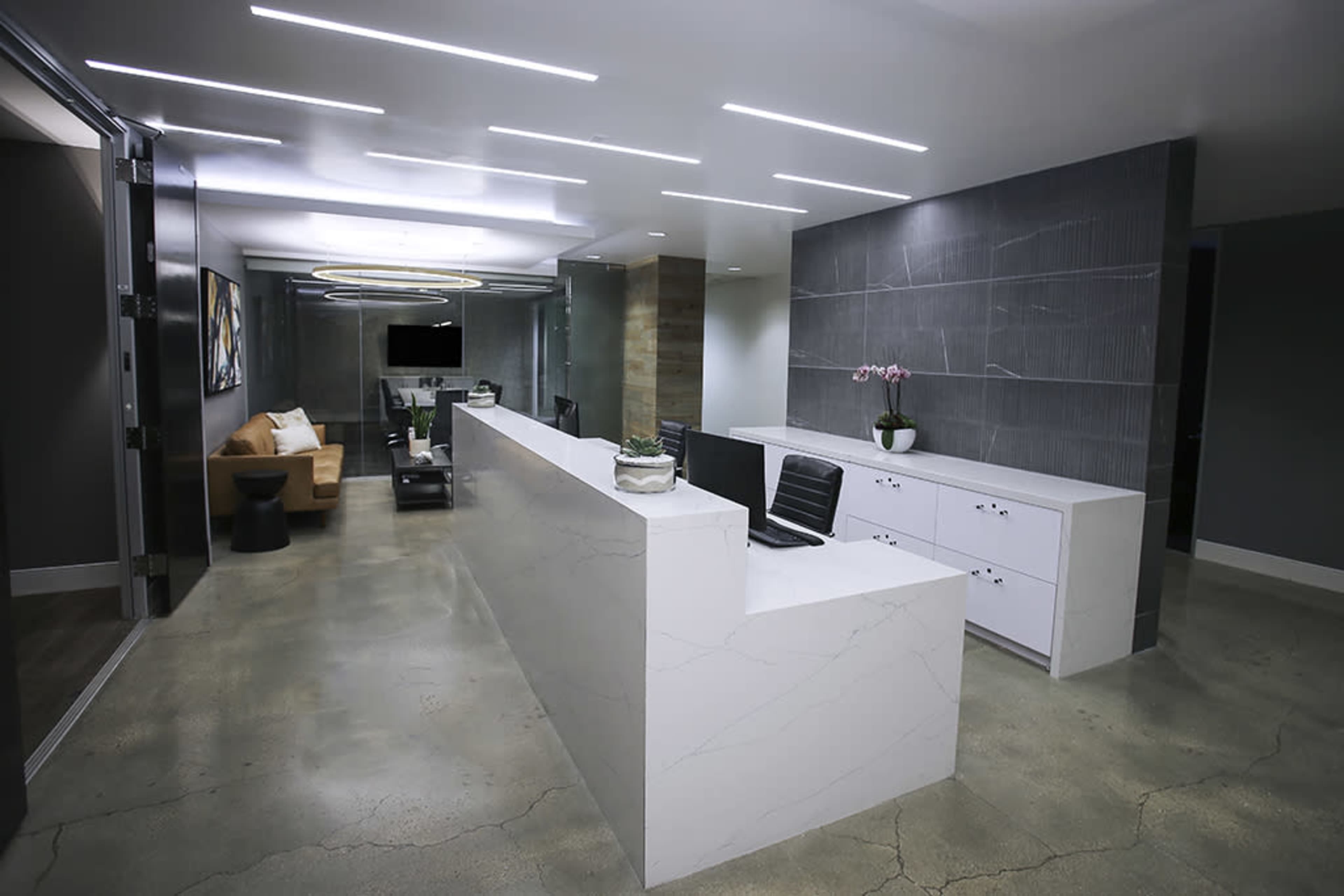 The image shows a modern office reception area with a white marble front desk, a seating area with a couch, and contemporary lighting fixtures.