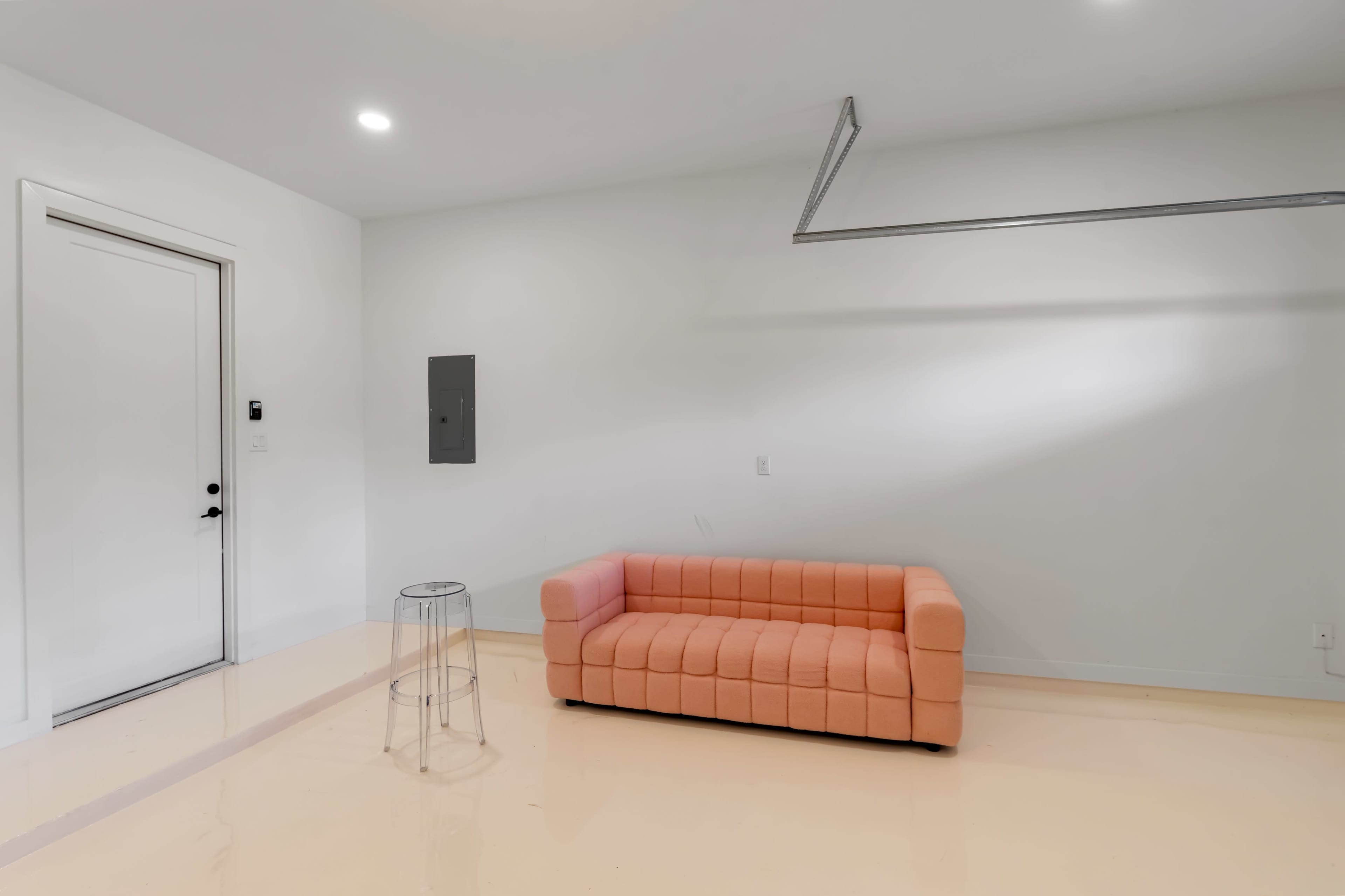 The image shows a minimalist interior with a light-colored floor, an orange sofa, a transparent side table, and a door on the left side.