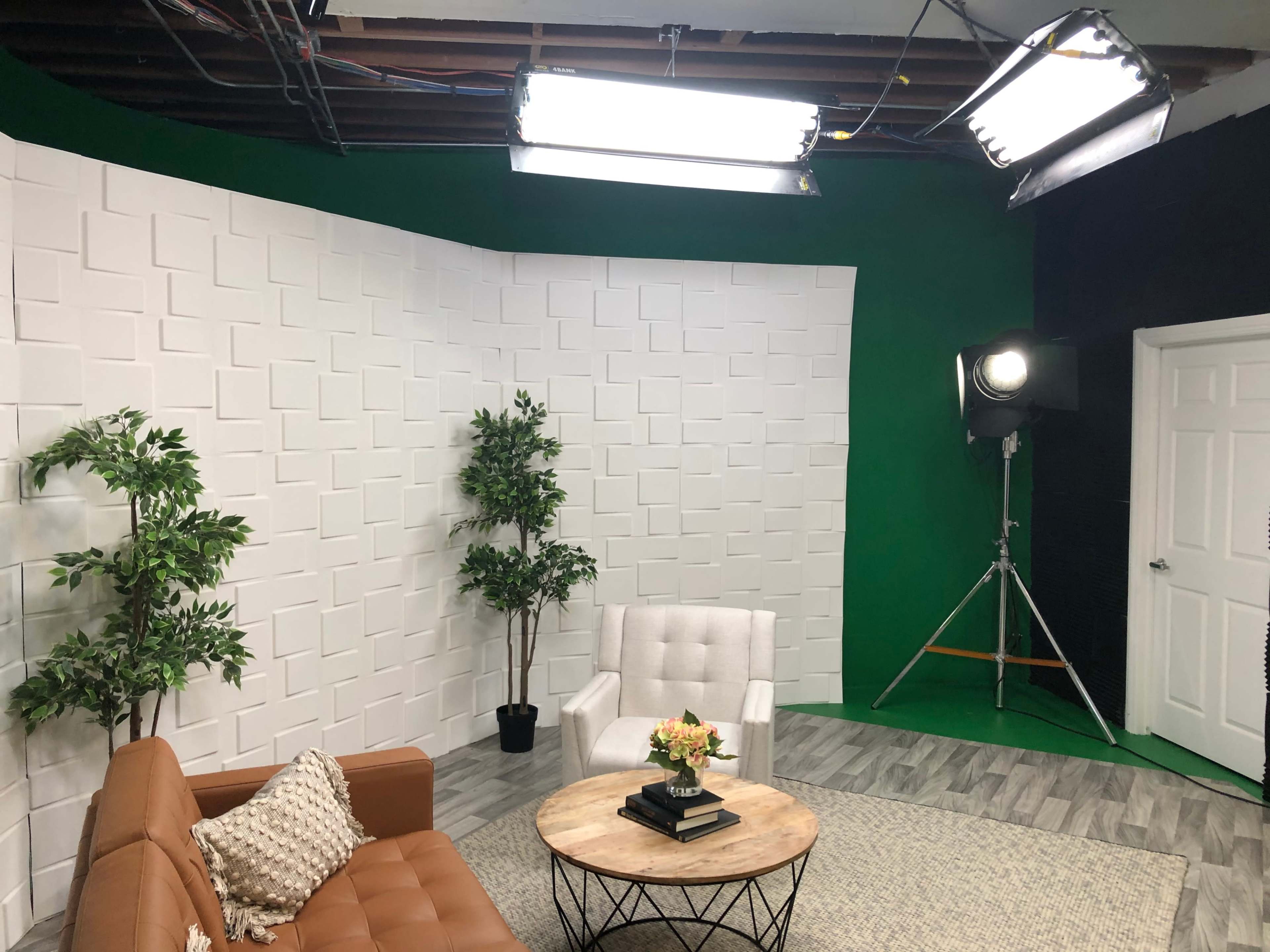 A studio setting features a brown sofa, a white armchair, a round coffee table with flowers, and green and black backdrop walls.