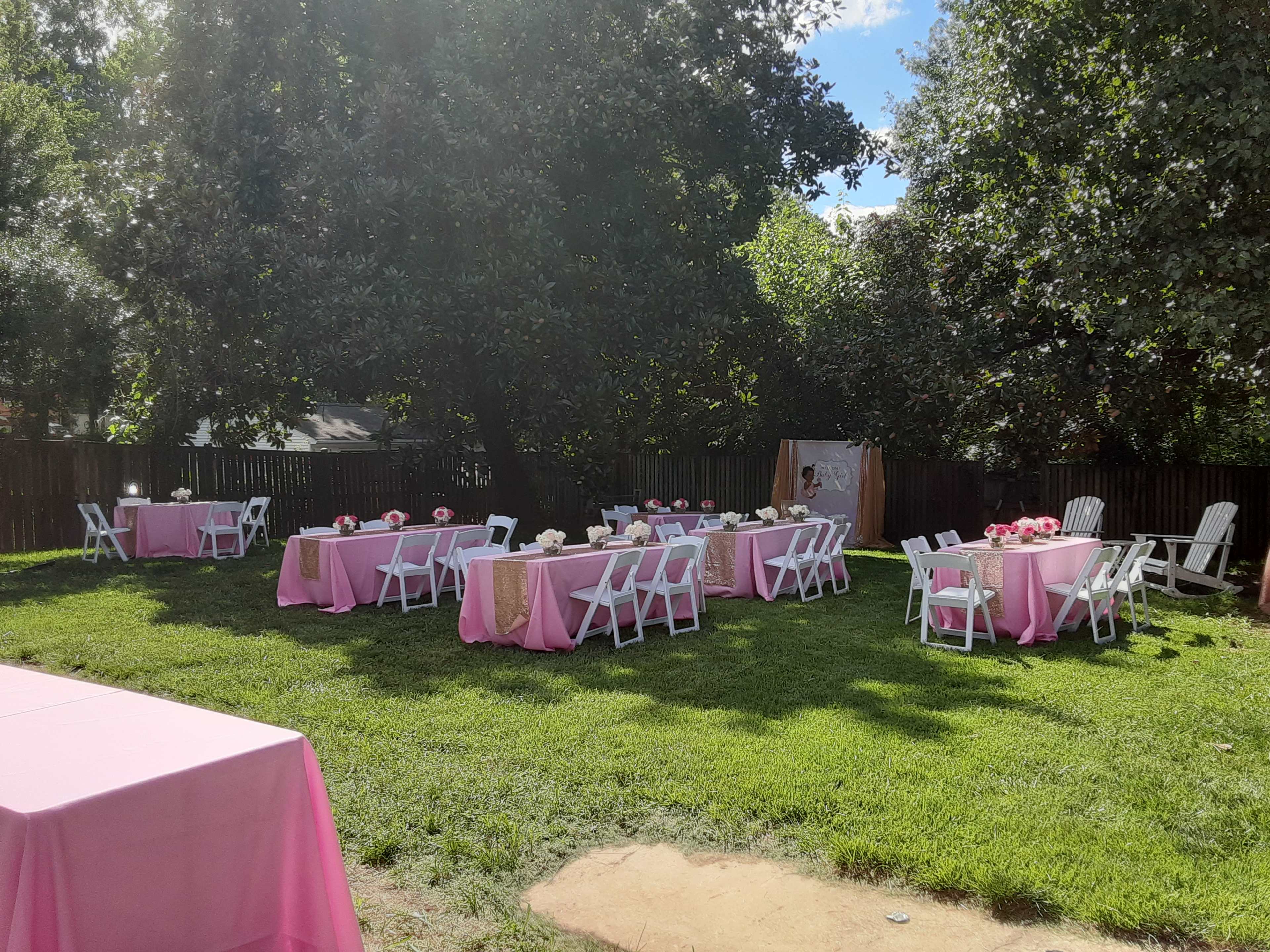 The image shows a backyard set up for an outdoor event, featuring several tables covered with pink tablecloths and surrounded by white chairs, all situated on green grass.