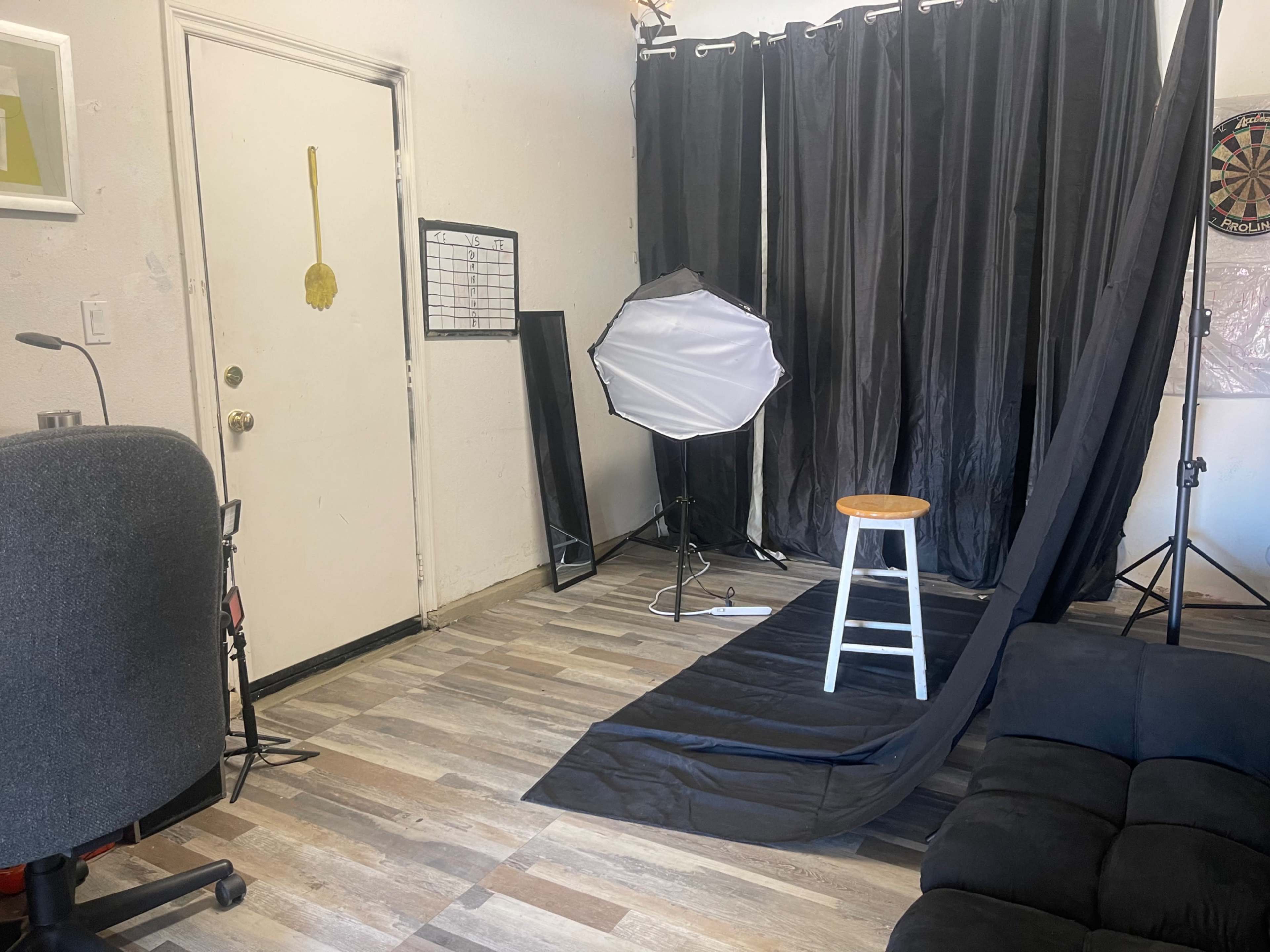 A small photography studio features a white stool on a black backdrop, with lighting equipment and a dartboard visible in the background.