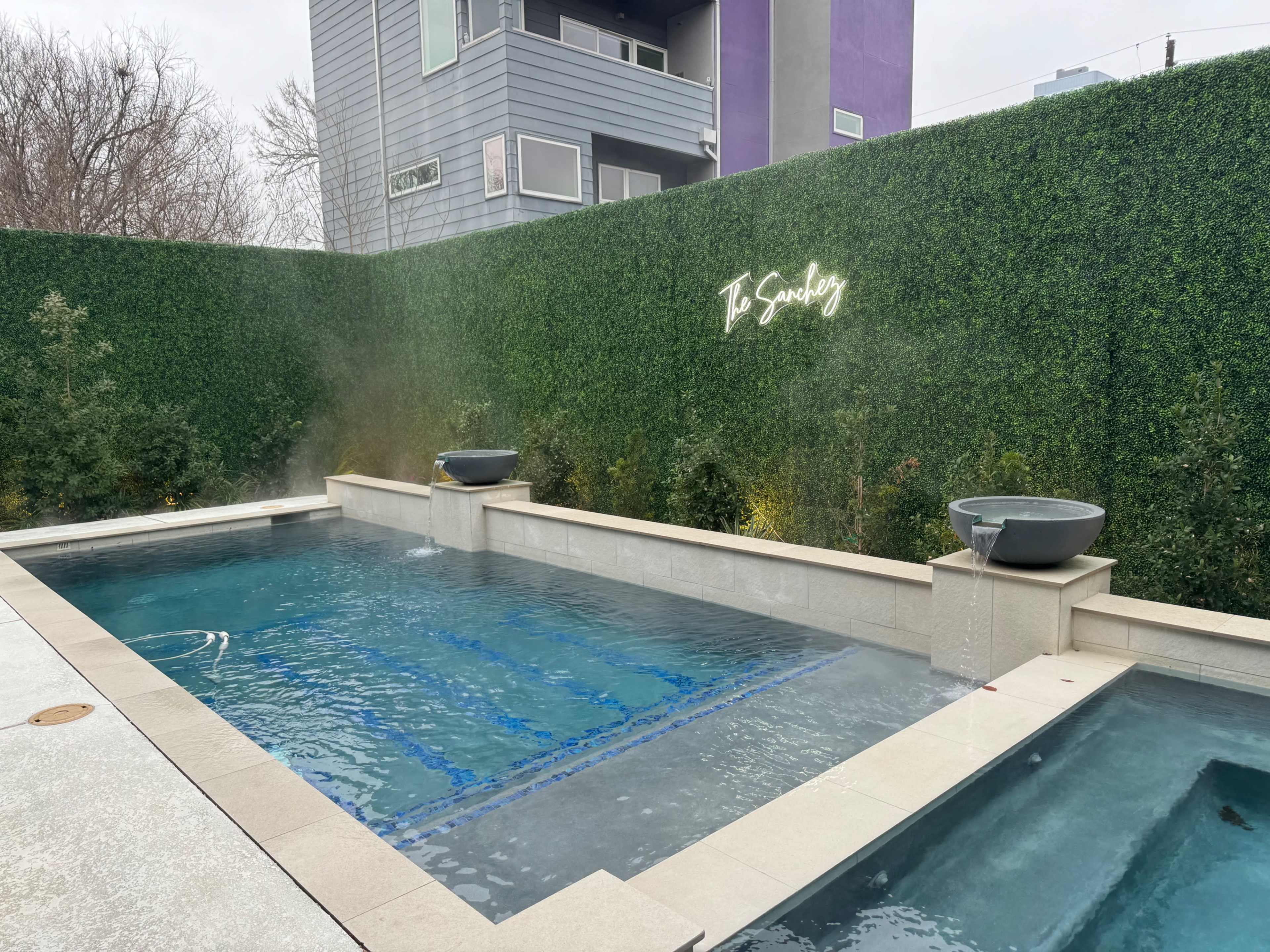 A modern swimming pool surrounded by a green hedge and features two water bowls at either end.